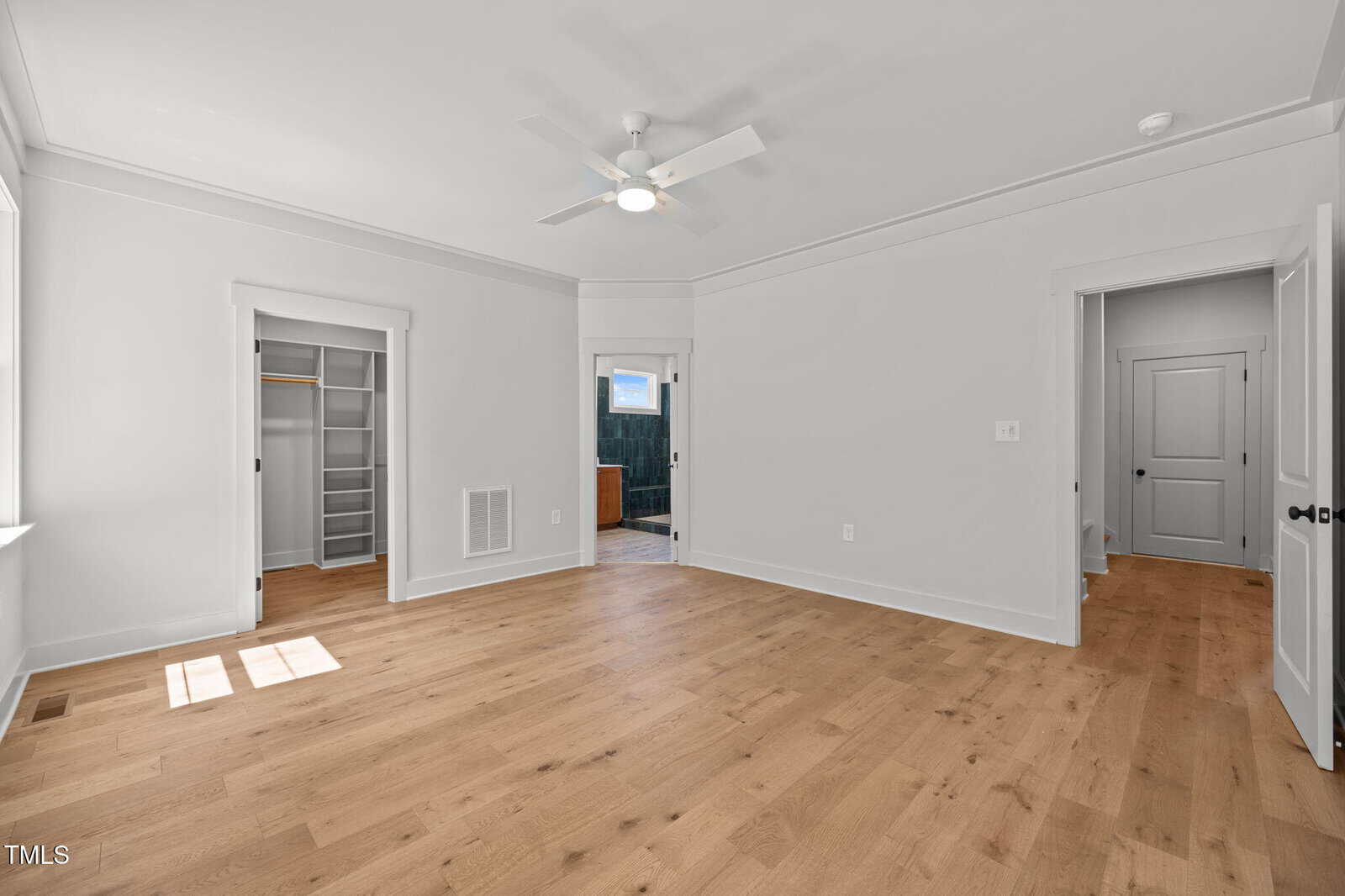Bright, empty bedroom with light wood floors, white walls, a ceiling fan, closet with shelving, en suite bathroom, and multiple white doors. Sunlight streams through a window on the left.