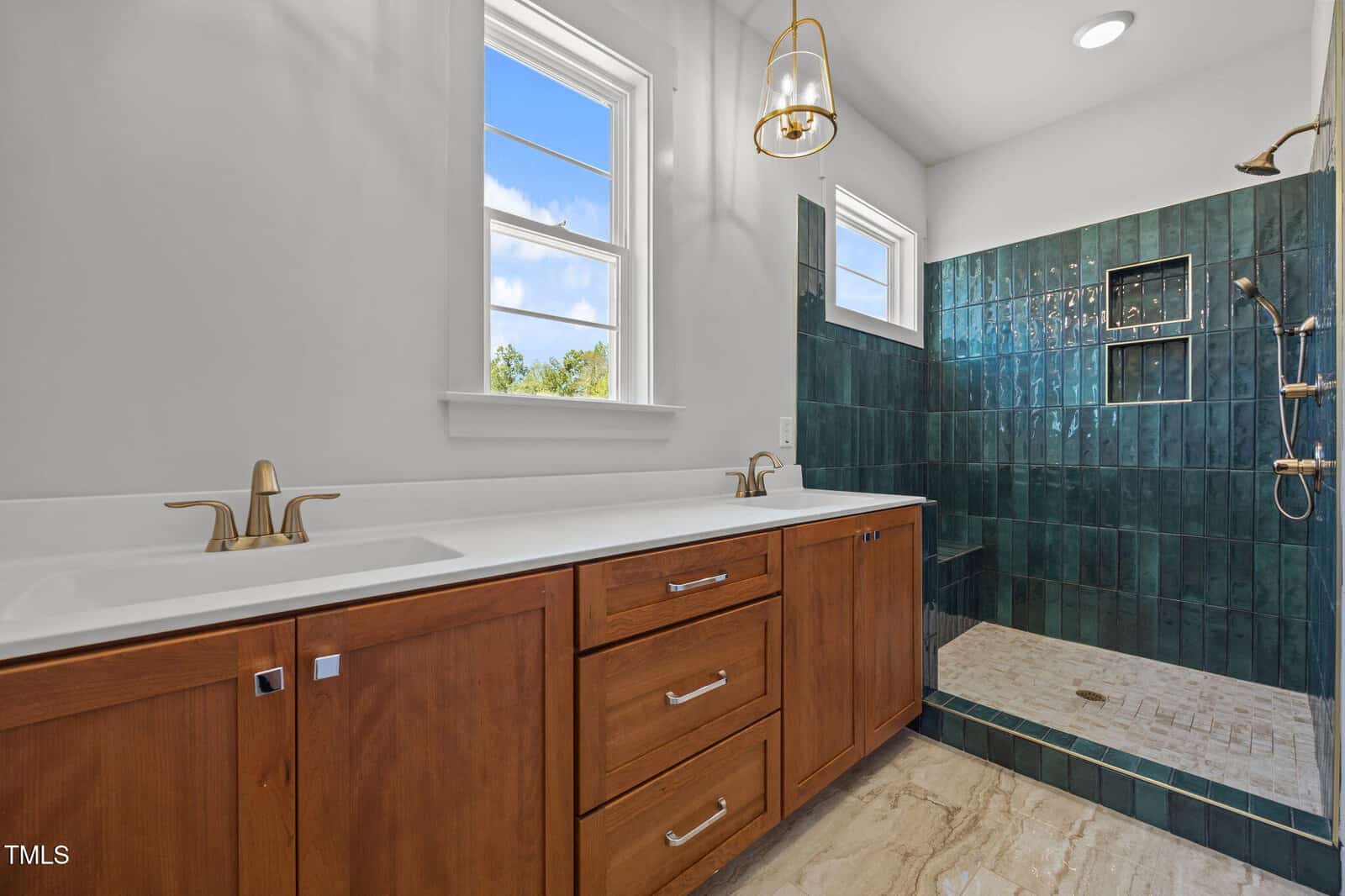 Modern bathroom with double sinks, wood cabinets, gold fixtures, and a walk-in shower with dark green tiles and built-in shelves. Two windows provide natural light.