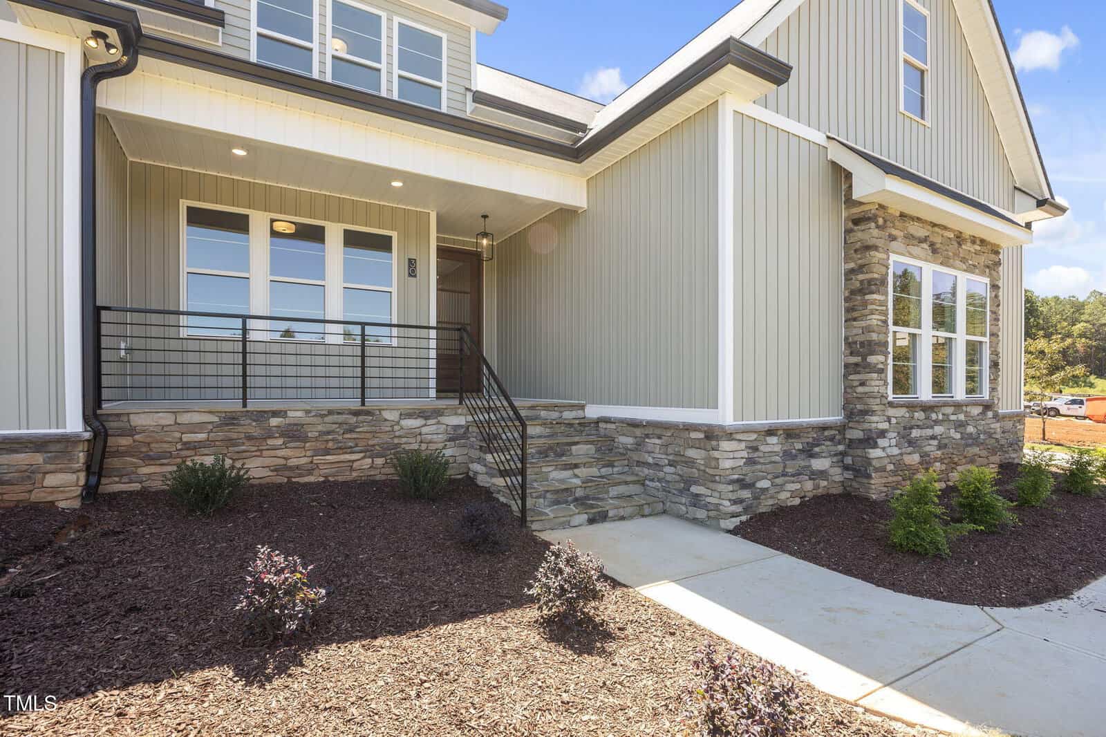 A modern house exterior with gray vertical siding, stone accents, a covered front porch with black railing, large windows, and landscaped shrubs along a concrete walkway.