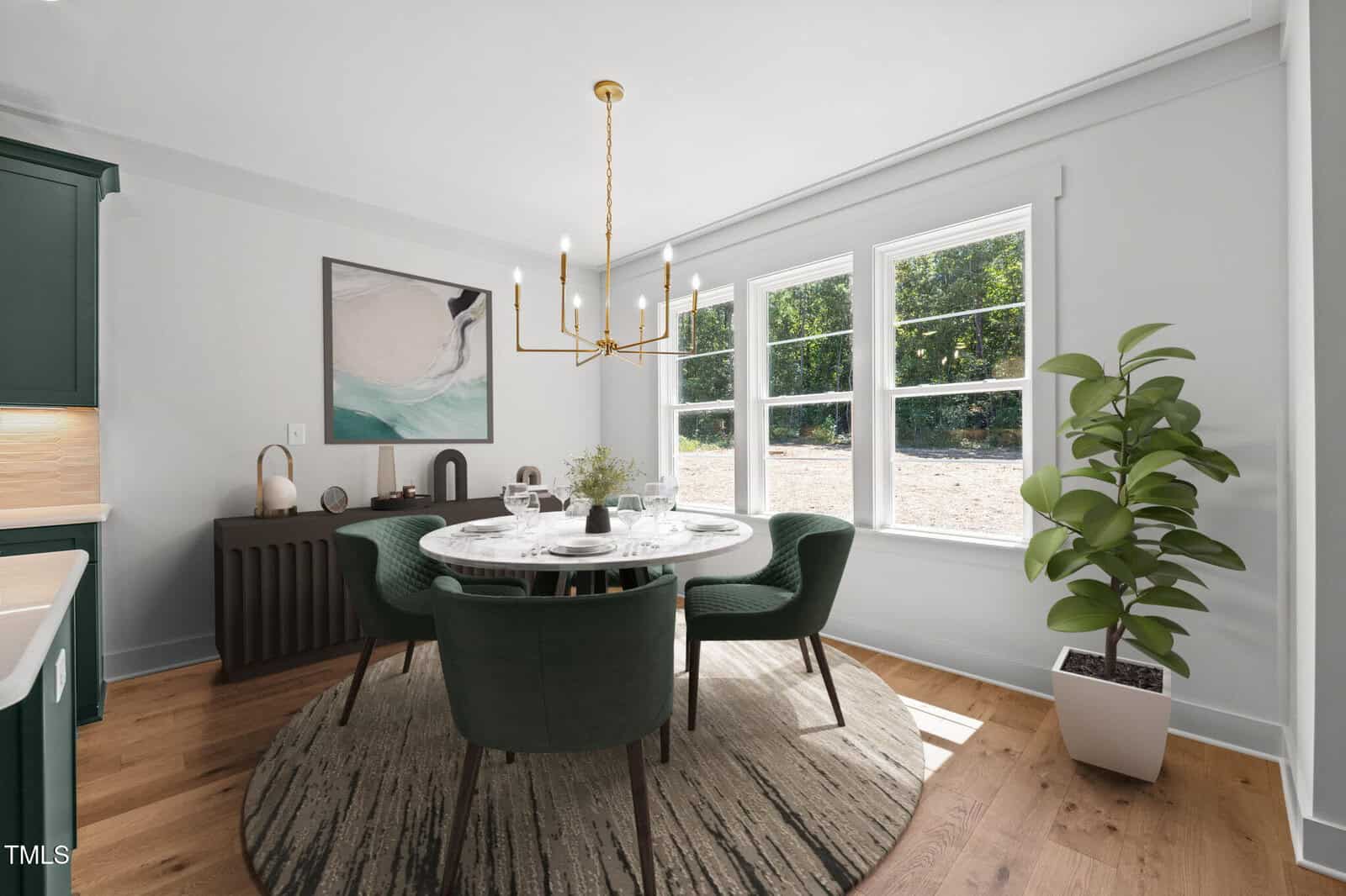 A modern dining room with a round table, four green chairs, a circular rug, potted plant, wall art, and large windows letting in natural light. A gold chandelier hangs above the table.