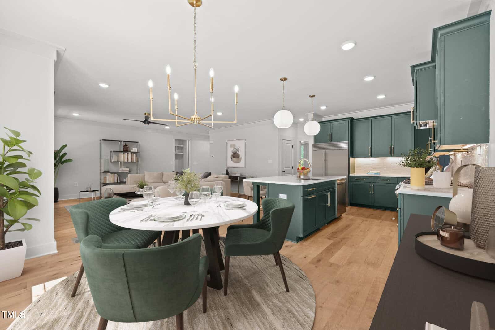 Modern open-plan kitchen and dining area with green cabinets, gold hardware, a round dining table set for four, large indoor plant, and cozy living space in the background with neutral decor.