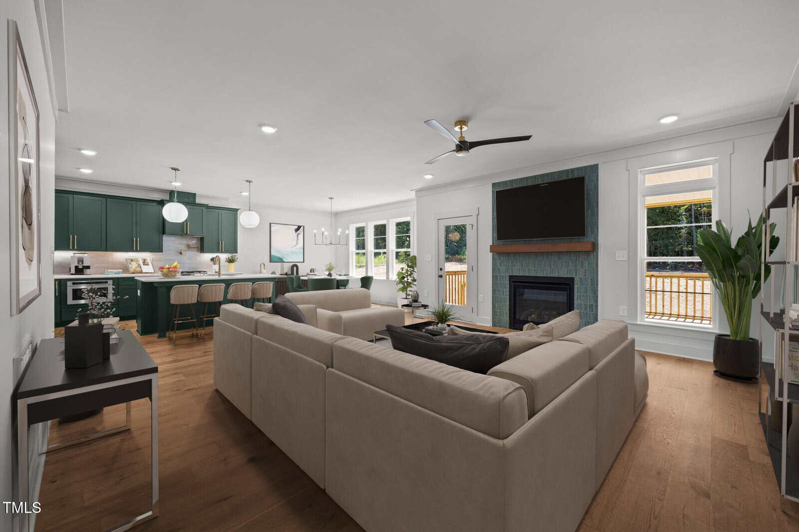 Open-concept living space with a beige sectional sofa, wall-mounted TV above a green-tiled fireplace, large kitchen with green cabinets and island seating, wood floors, and large windows letting in natural light.
