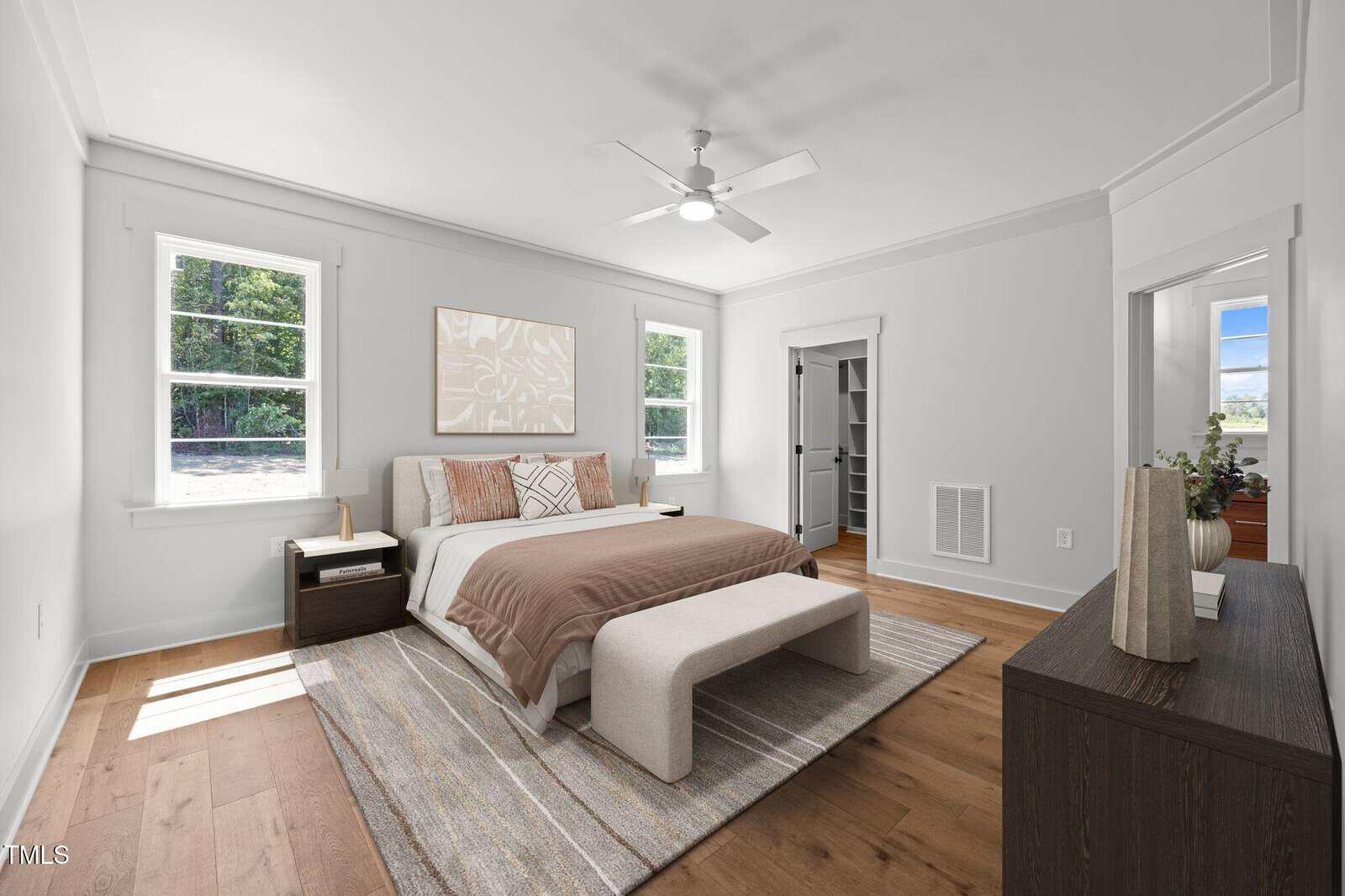 A bright bedroom with a large bed, beige bedding, two windows, a bench, nightstands, a dresser with decor, wood floors, a ceiling fan, and an open door to a walk-in closet.