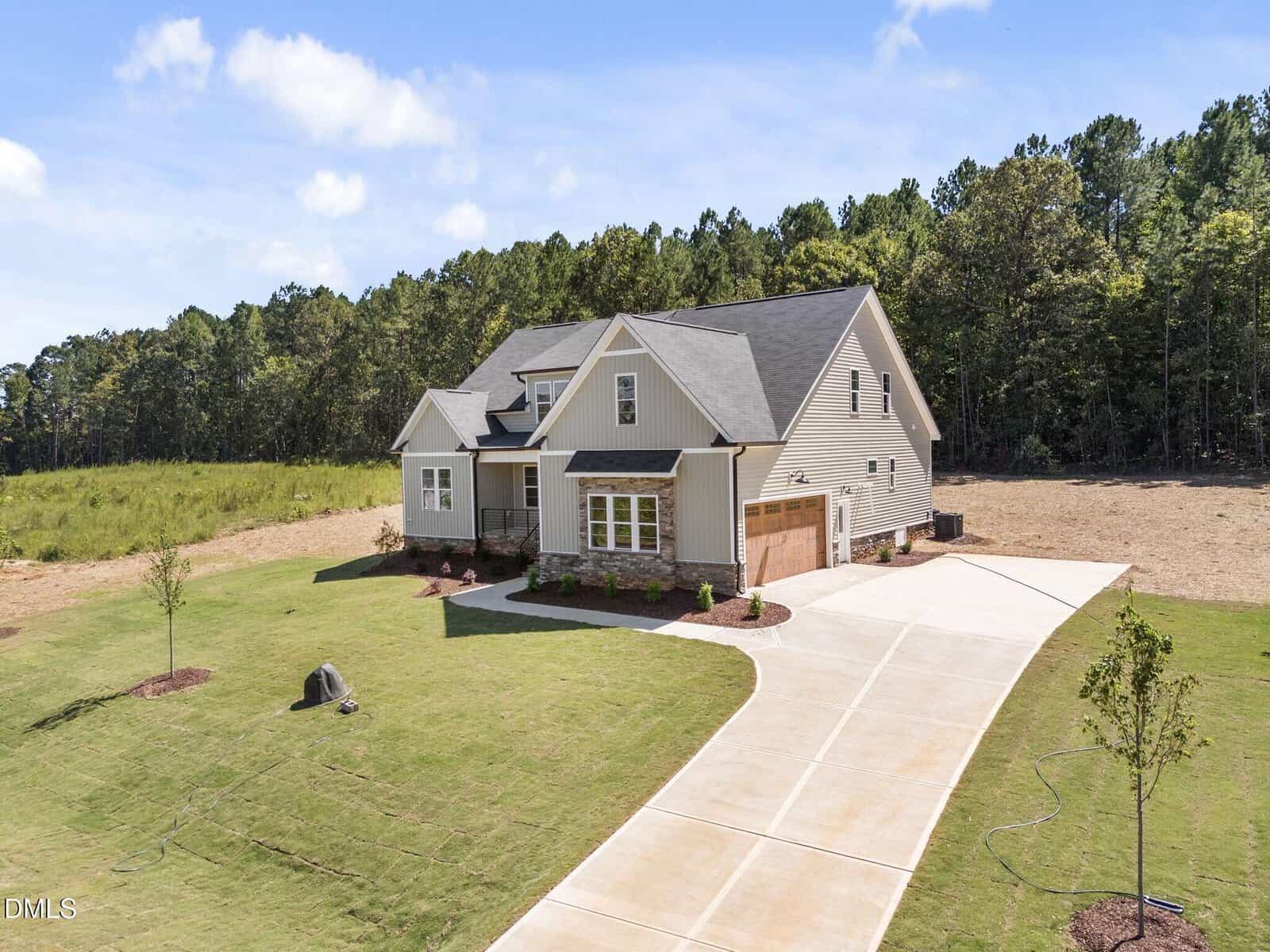 A modern suburban house with beige siding, a gabled roof, and a two-car garage sits on a spacious lot with a large driveway, young trees, and a grassy lawn, bordered by woods under a blue sky.