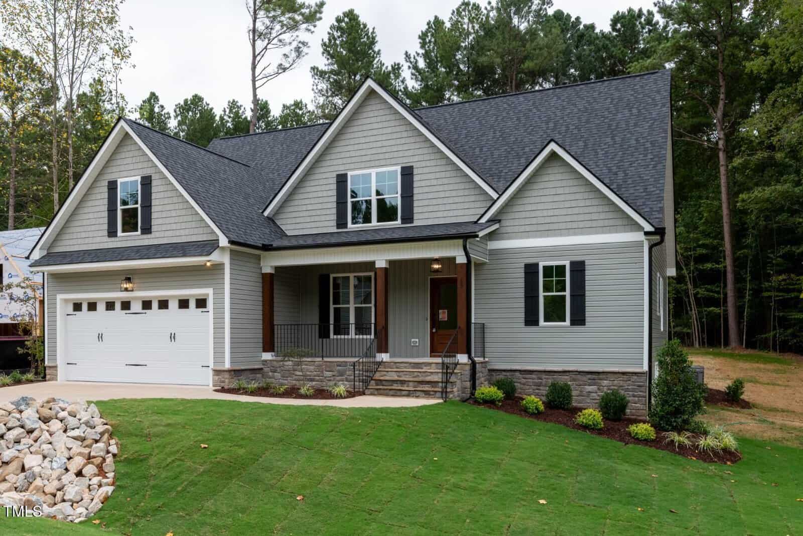 A modern two-story house with gray siding, white trim, a black roof, and a covered front porch. The front yard features green grass, landscaped bushes, and a stone-lined path leading to a white garage door.