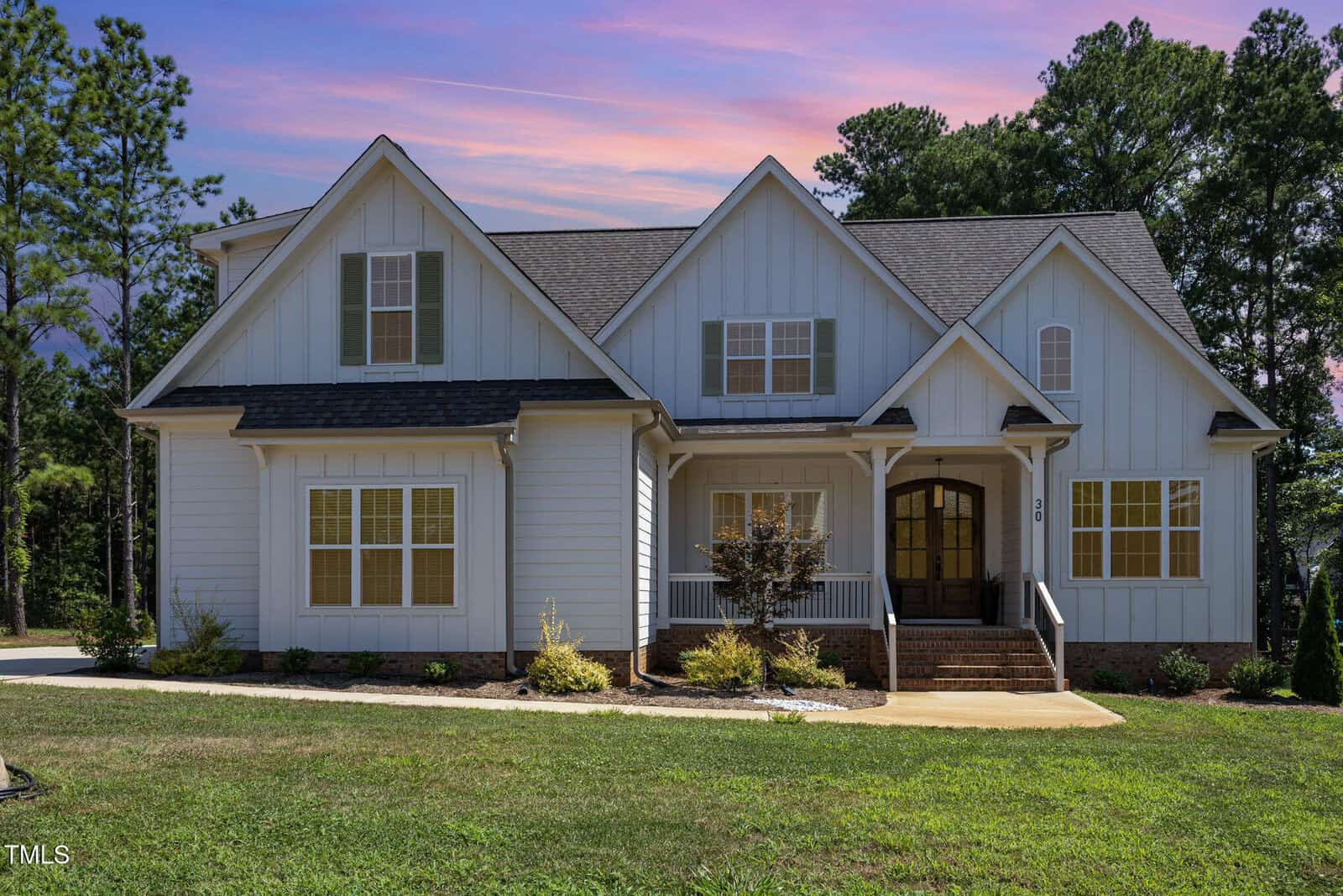 A white, two-story house with gray roof shingles, board-and-batten siding, double front doors, front steps, and a small landscaped yard sits at 30 Hamilton Way in Youngsville, set against a backdrop of trees and a colorful evening sky.