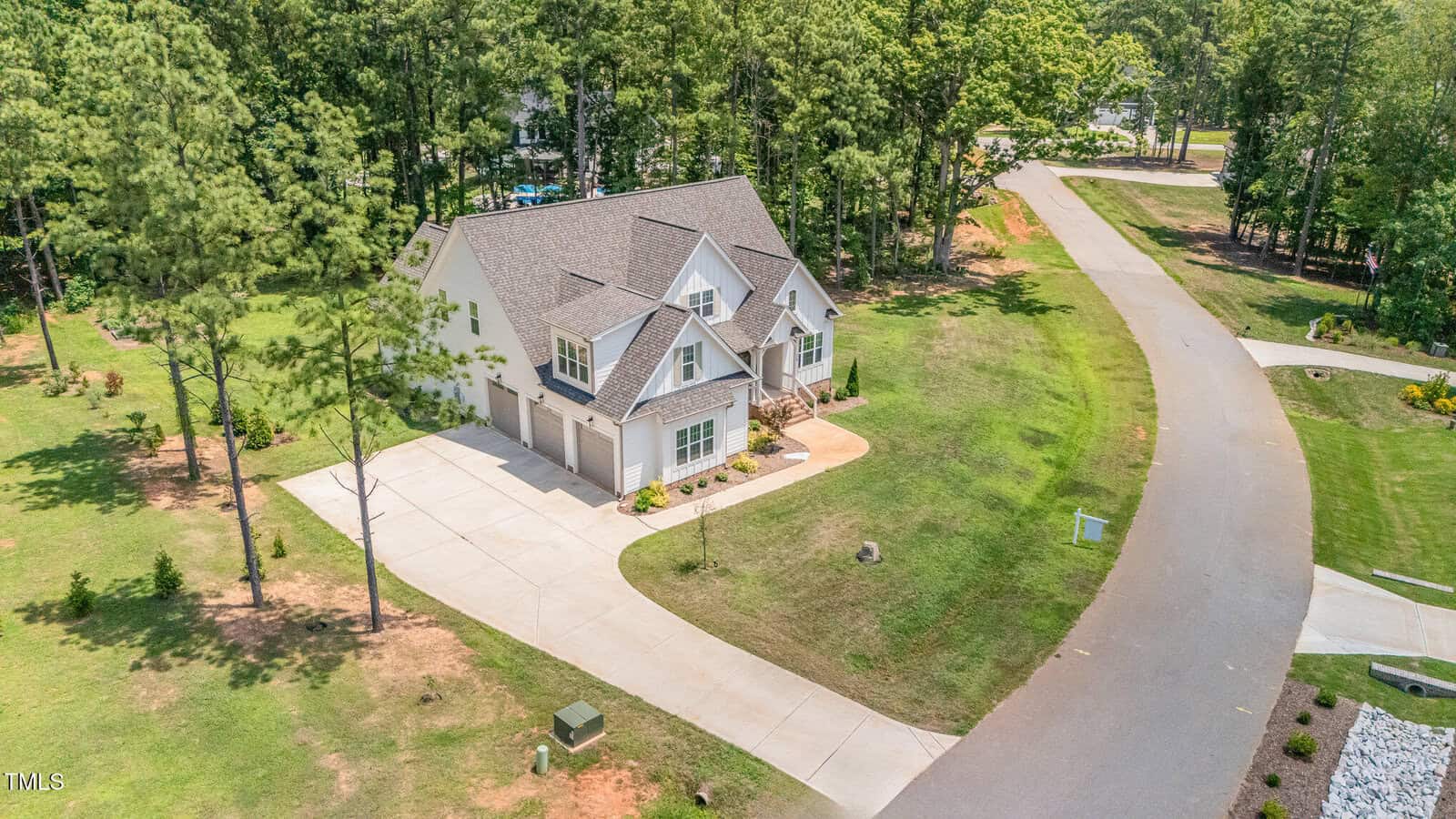 Aerial view of a two-story suburban house with a three-car garage, surrounded by lawns and trees, located at a corner of a curved, paved street in a residential neighborhood.