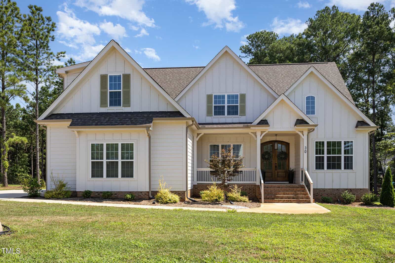 A two-story white house with board and batten siding, multiple peaked roofs, and brick steps leading to a double front door. The house sits on a grassy lawn with small shrubs and trees, backed by tall pine trees.