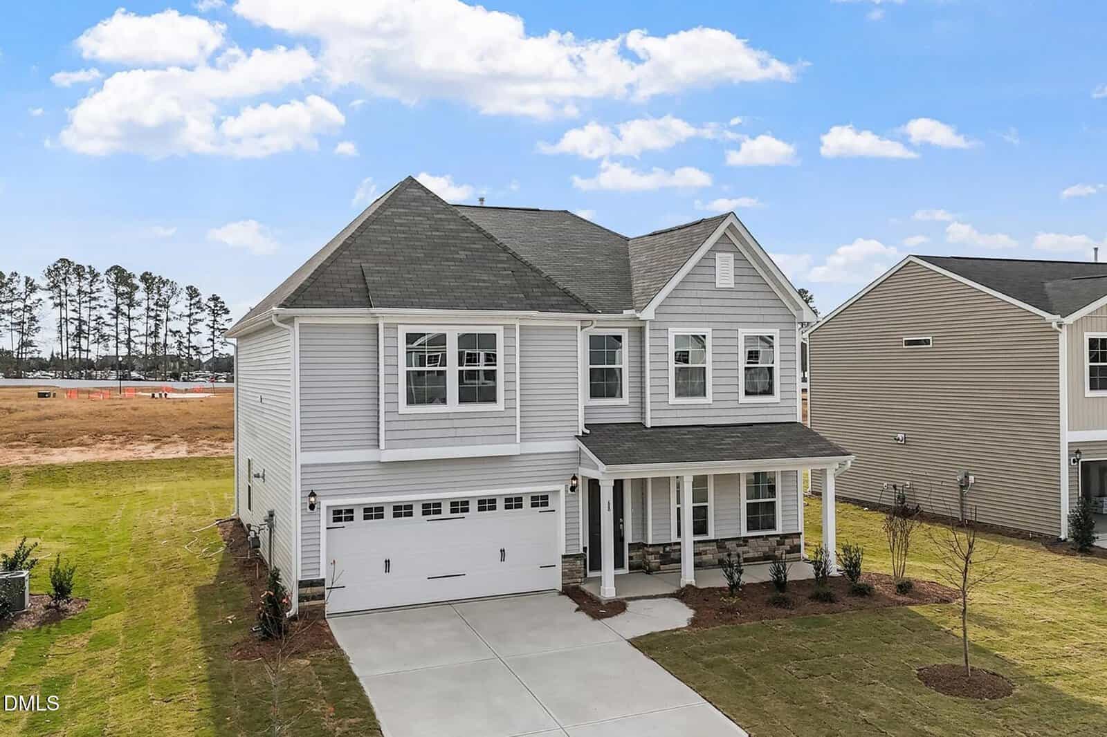 Two-story suburban house with gray siding, white trim, and a covered front porch. Attached two-car garage with white doors. The yard is landscaped with grass and small plants. Blue sky with scattered clouds in the background.