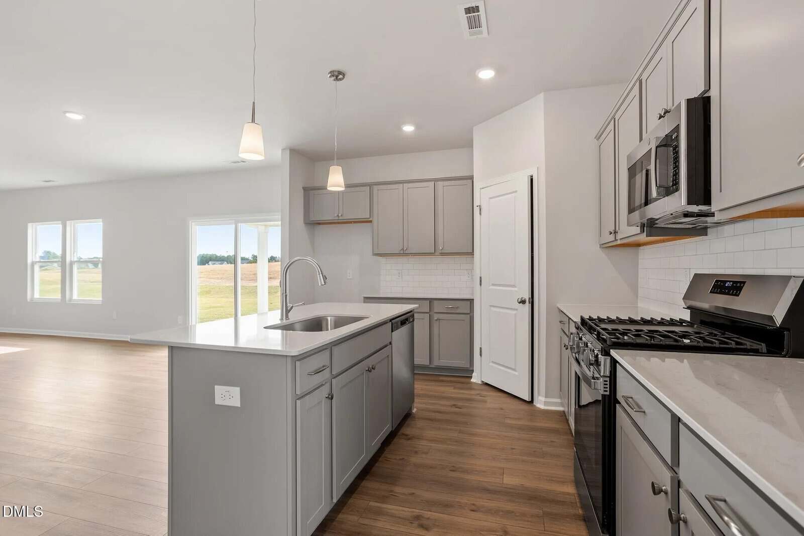 Modern kitchen with light gray cabinets, stainless steel appliances, white tiled backsplash, and an island with a sink. The room has wood flooring, hanging pendant lights, and an open layout connected to a bright living area.