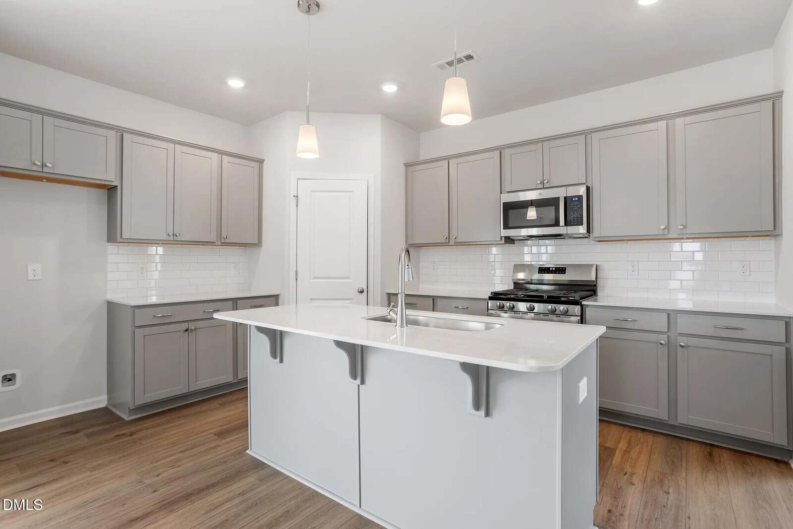 Modern kitchen with gray cabinets, white countertops, an island with a sink, stainless steel appliances, subway tile backsplash, pendant lights, and wood flooring. Walls are light-colored, and there is a white door at the back.