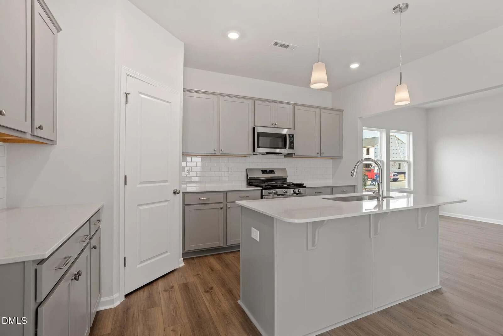 Modern kitchen with light gray cabinets, white countertops, a central island with pendant lights, stainless steel appliances, white subway tile backsplash, wood flooring, and a pantry door on the left.