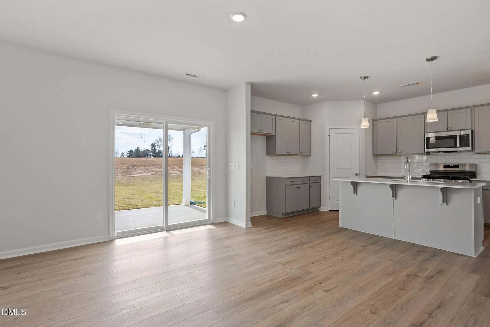 Modern kitchen and living area with light wood flooring, gray cabinets, stainless steel appliances, white countertops, and pendant lights. Sliding glass doors lead to a backyard with a grassy open area and a covered patio.