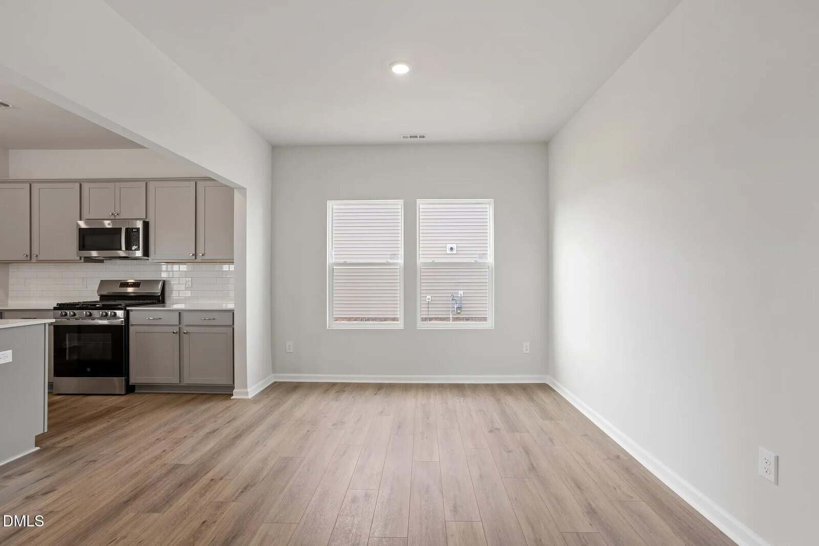 A modern, unfurnished kitchen and dining area with light wood flooring, gray cabinets, stainless steel appliances, white subway tile backsplash, and two windows on the back wall letting in natural light.