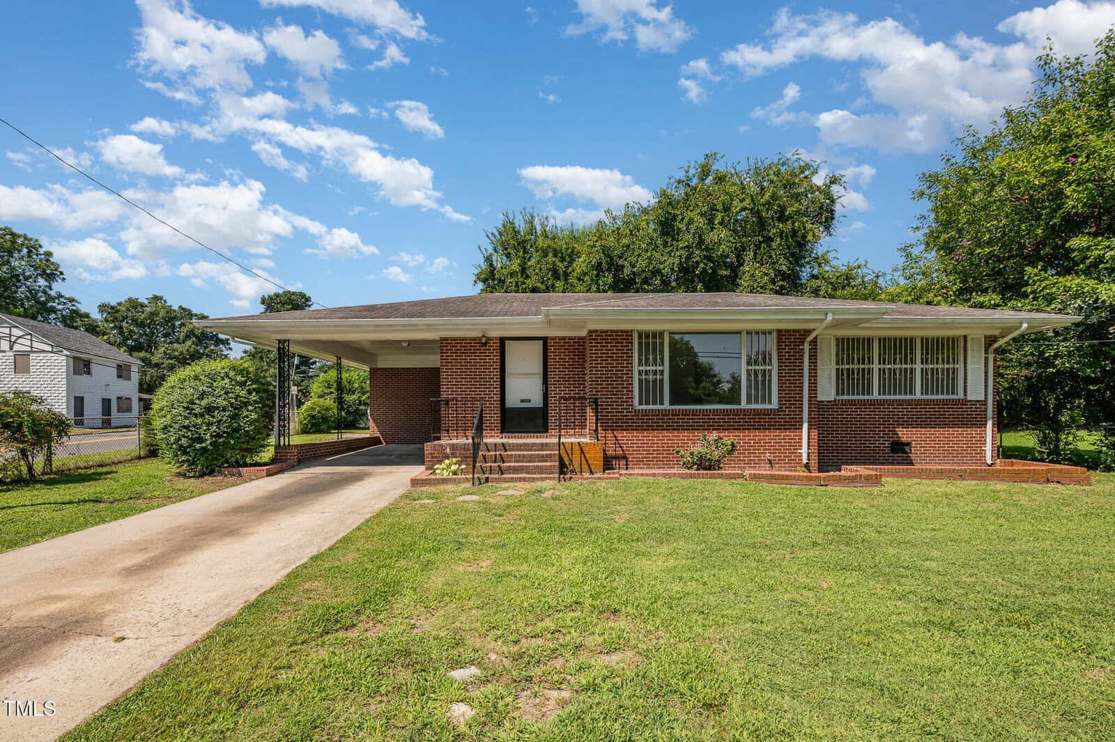 A single-story brick house with a carport on the left, a concrete driveway, front steps, a white door, large windows with white bars, and a grassy front yard under a blue sky with scattered clouds.