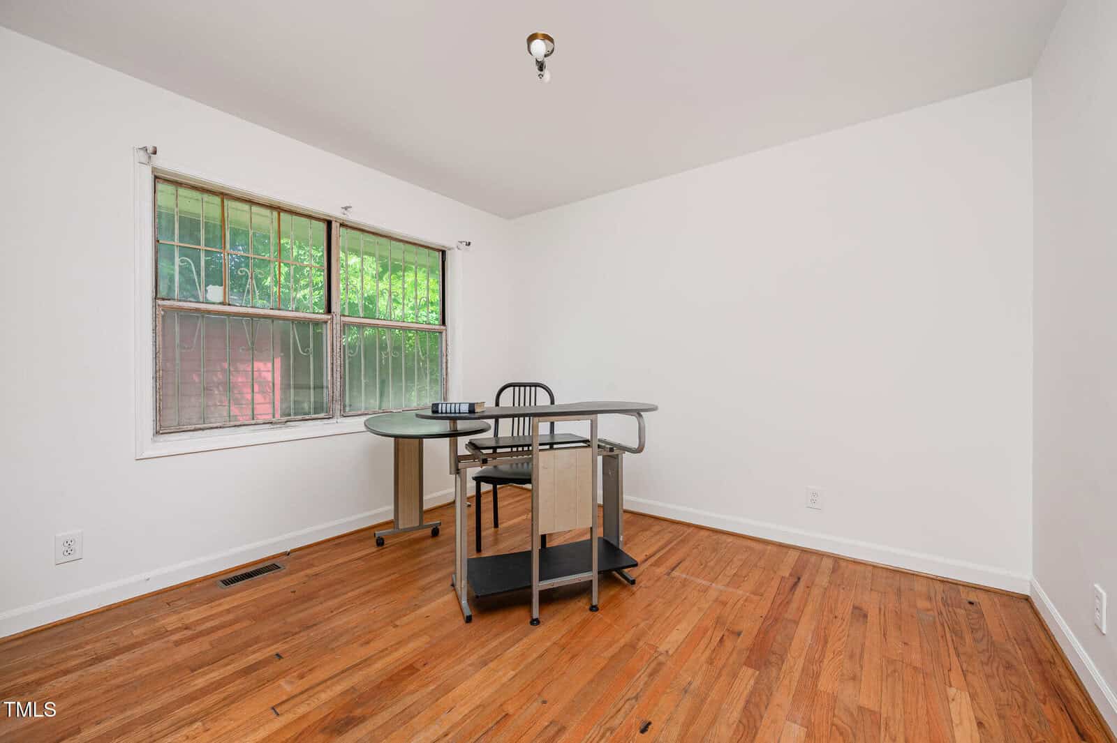 A mostly empty room with hardwood floors, white walls, a single window with security bars, and a corner desk with a chair. The ceiling has an exposed light fixture without a cover.