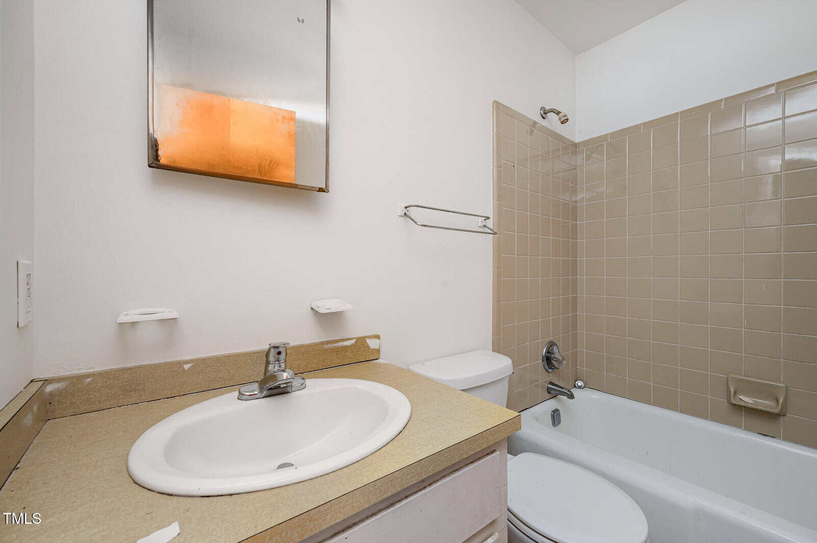 A bathroom with a beige countertop, white sink, wall-mounted mirror, toilet, and a bathtub with beige tile surround. Two empty soap holders are above the sink, and a towel rack is on the wall next to the tub.