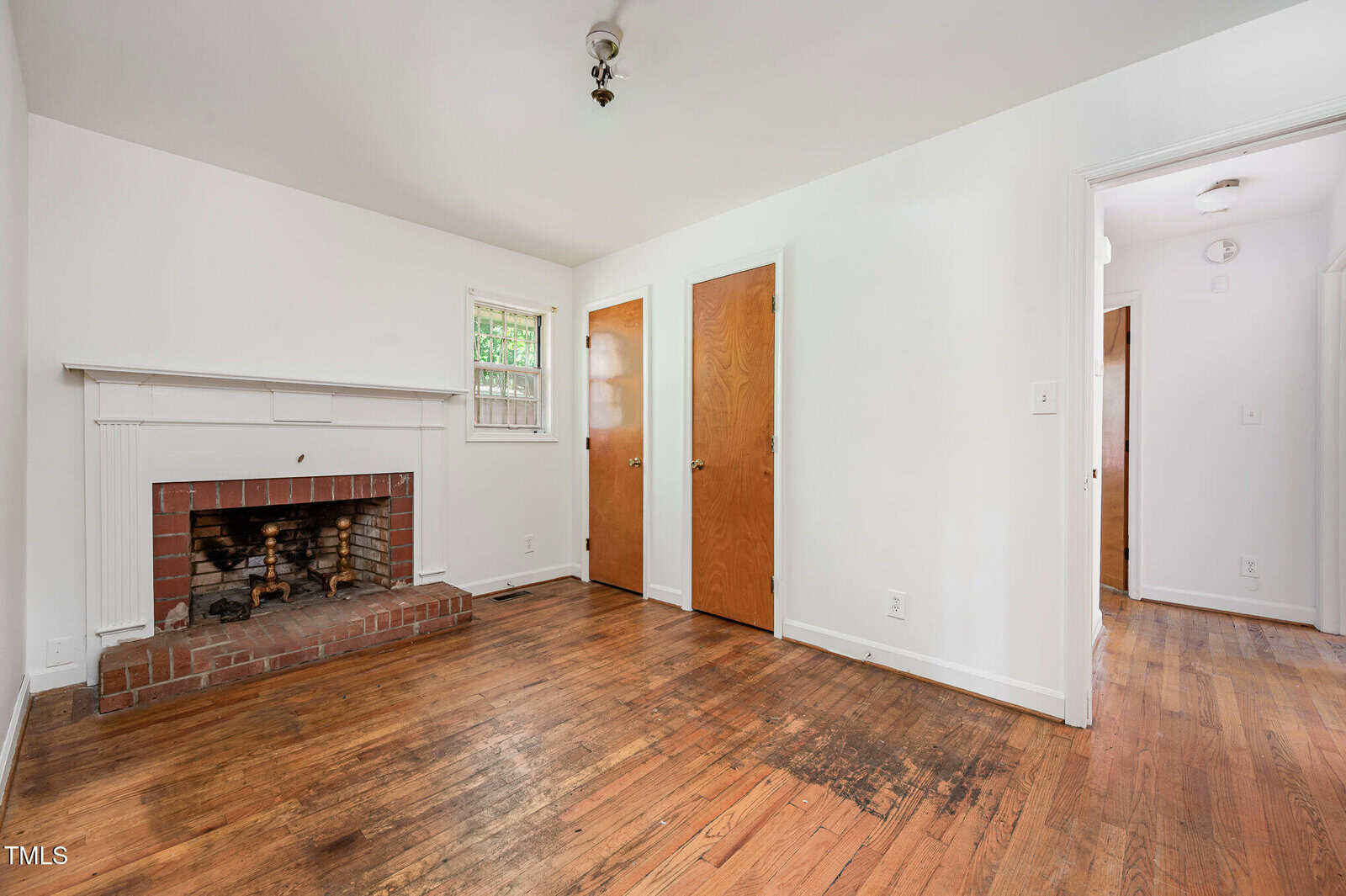 A small, empty room with white walls, a brick fireplace, hardwood floors, two wooden doors, a window, and a hallway entrance. The floor shows signs of wear and there is a light fixture on the ceiling.