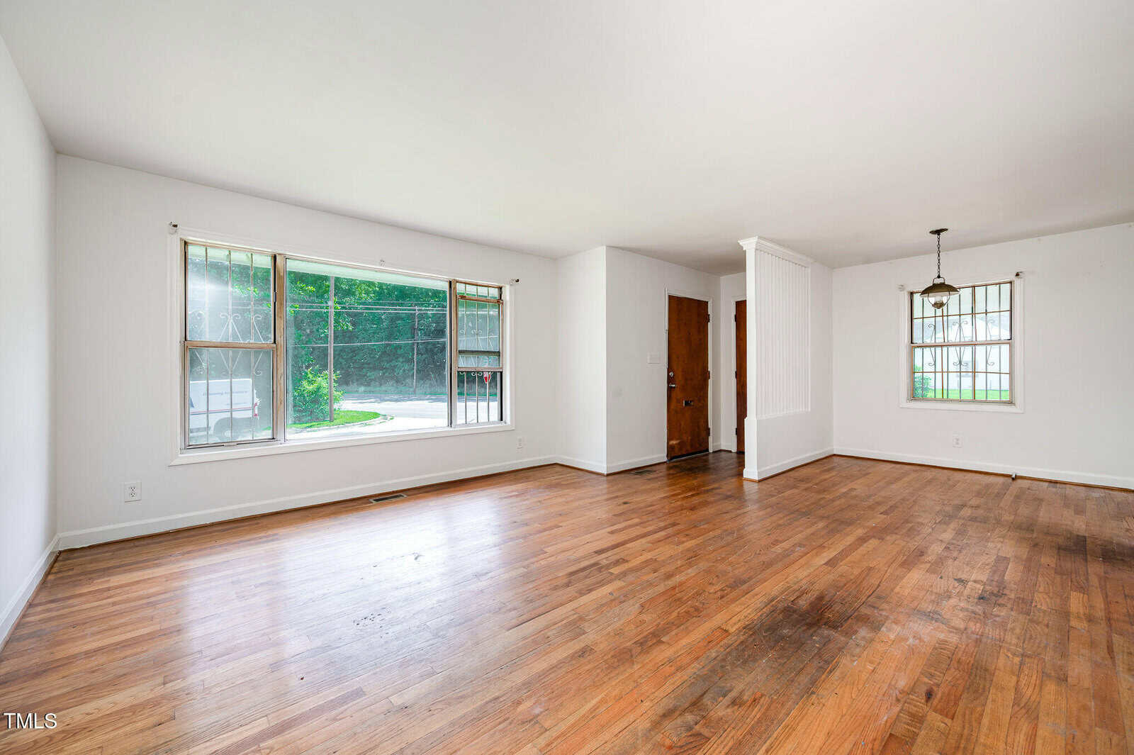 Unfurnished living and dining area with white walls, wood floors, two large windows, a hanging light fixture, and a wooden front door. The space is open and bright with natural light.