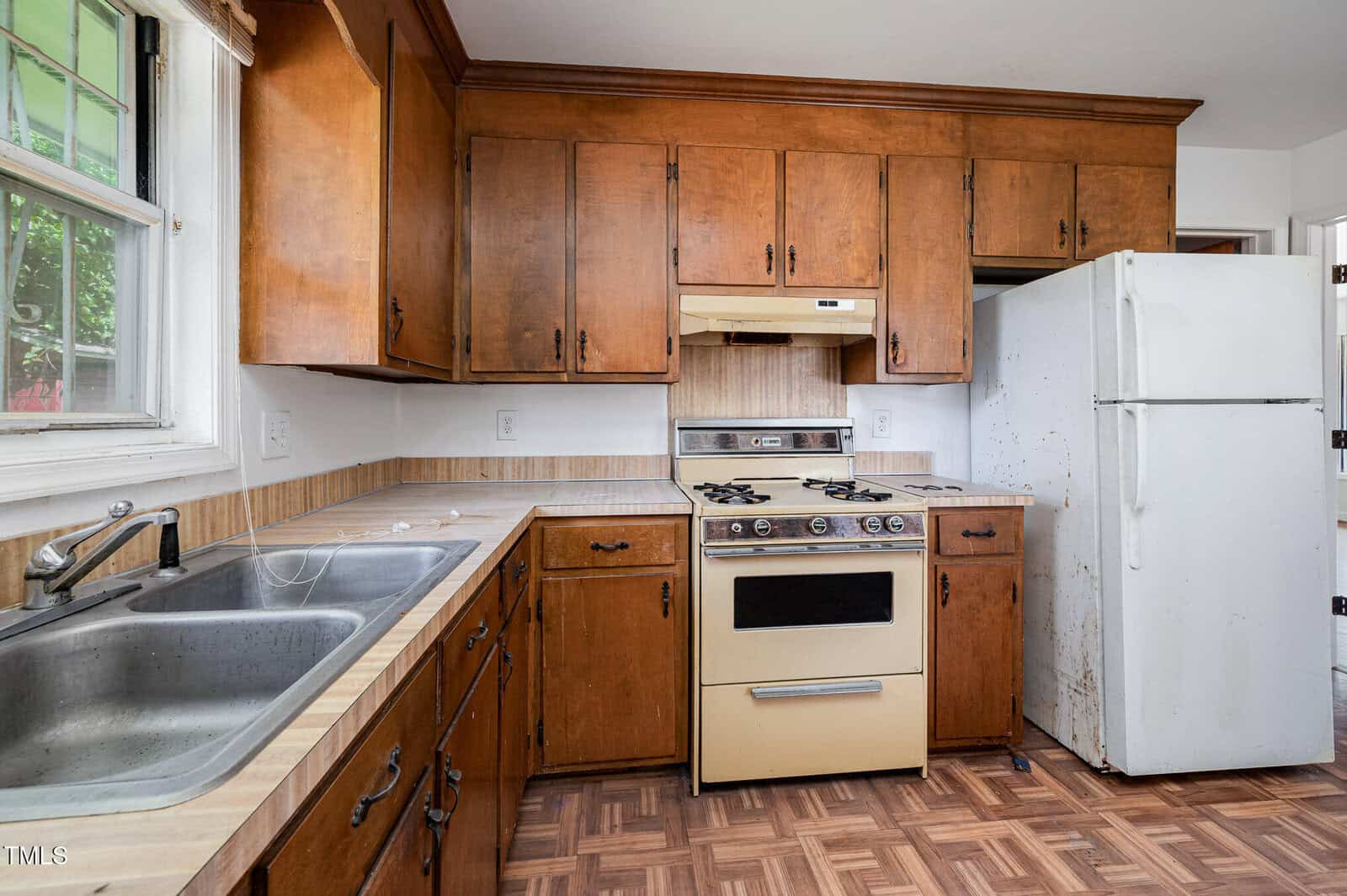 A kitchen with brown wooden cabinets, a white refrigerator with visible rust stains, a vintage gas stove with range hood, a double sink under a window, and laminate countertops with a wood-patterned floor.