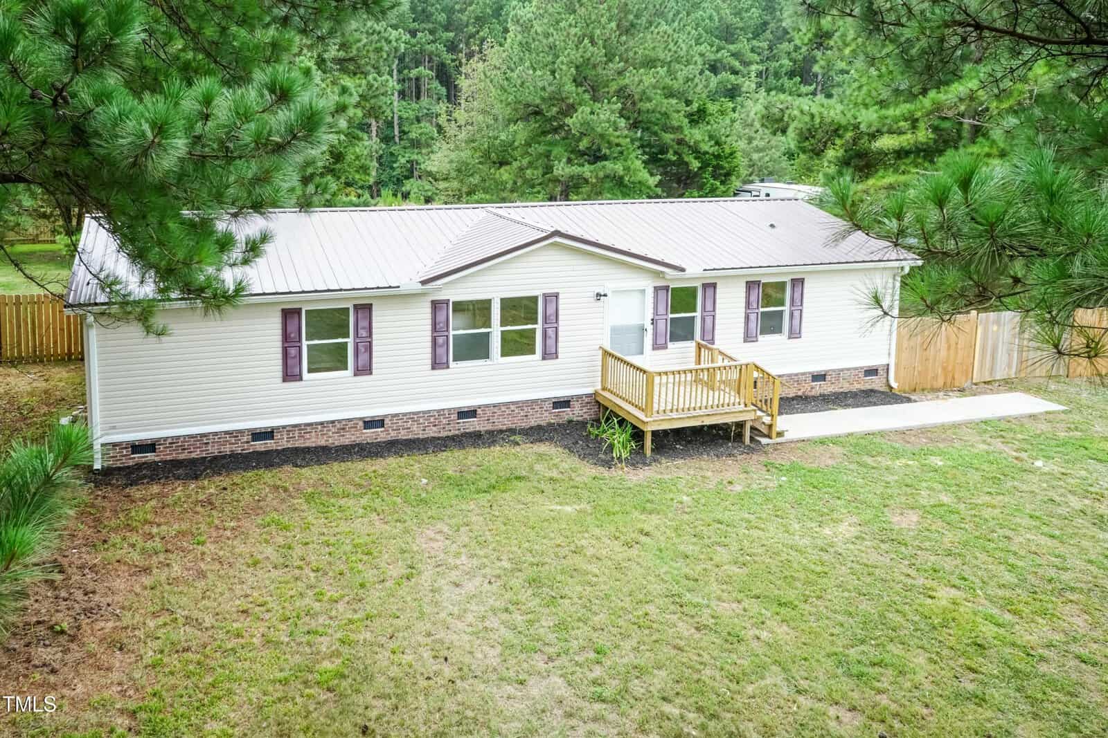 A single-story manufactured home with white siding, purple shutters, a metal roof, and a small wooden front porch; surrounded by grass and trees with a forested background.
