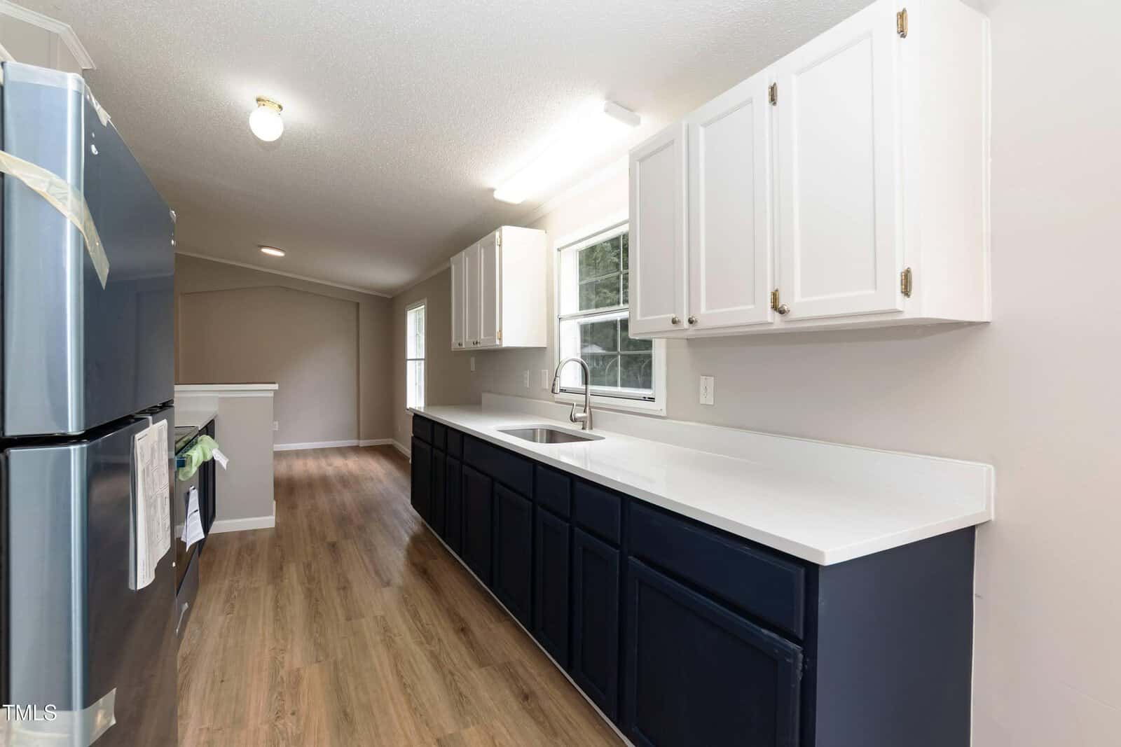 Modern kitchen with white upper and navy lower cabinets, white countertops, stainless steel refrigerator, wood flooring, and a large window above the sink, letting in natural light.