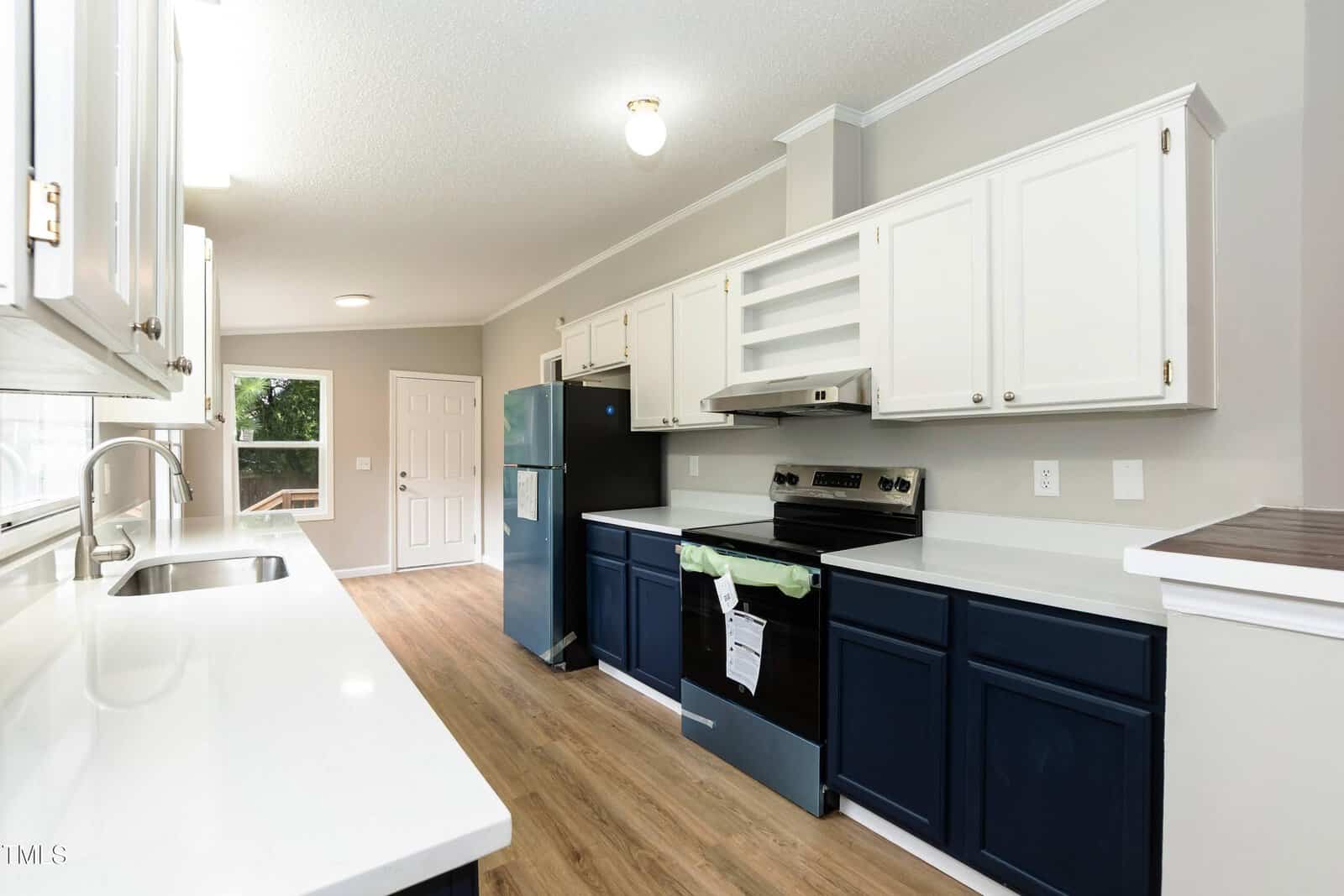 A modern galley kitchen with white upper cabinets, navy lower cabinets, white countertops, stainless steel appliances, wood flooring, and a view of a door and window at the far end.