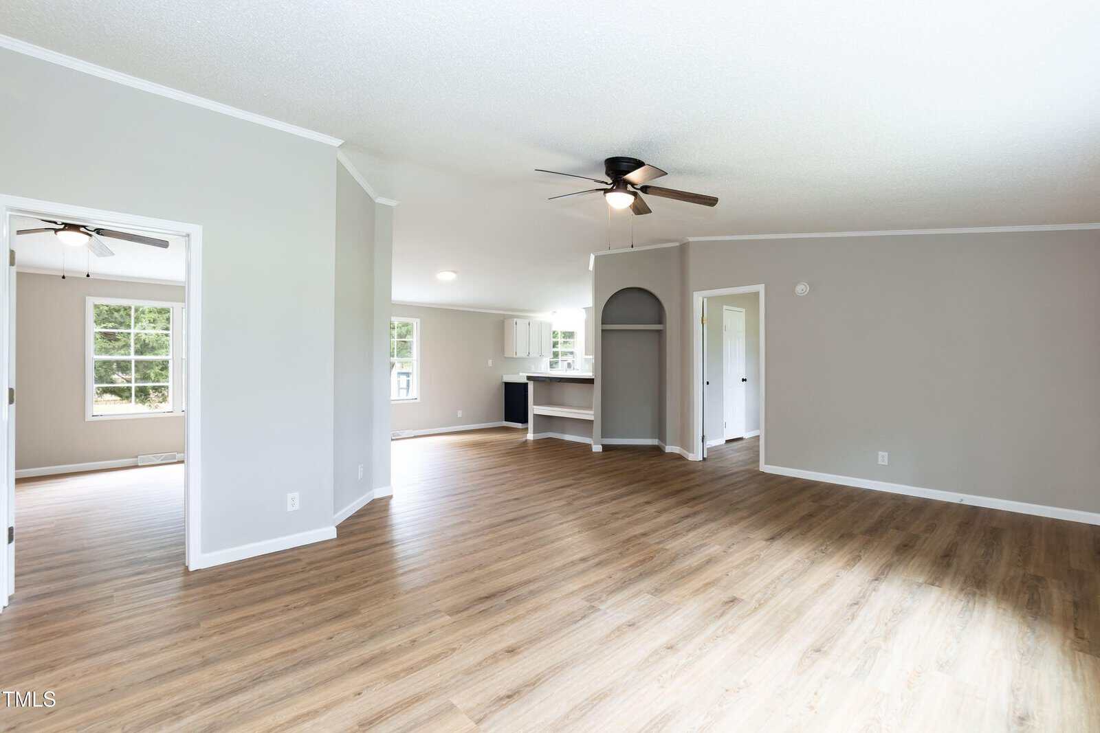 A spacious, empty living area with light wood flooring, light gray walls, a ceiling fan with lights, and large windows; a doorway reveals another room and an open kitchen is visible in the background.