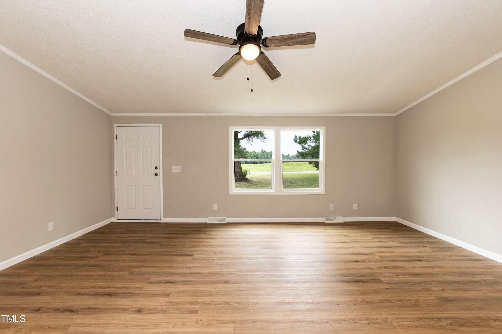 A spacious, empty living room with light beige walls, wood flooring, a ceiling fan with lights, a white front door, and two windows showing trees and grass outside.