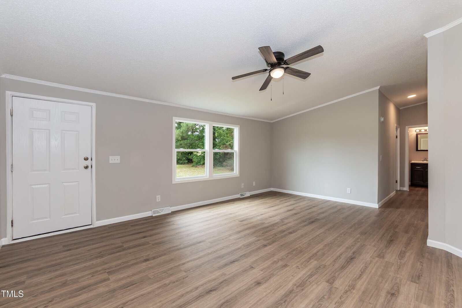A spacious, empty living room with light gray walls, wood flooring, a white door, two windows, ceiling fan with lights, and a hallway leading to a bathroom with a lit vanity mirror.