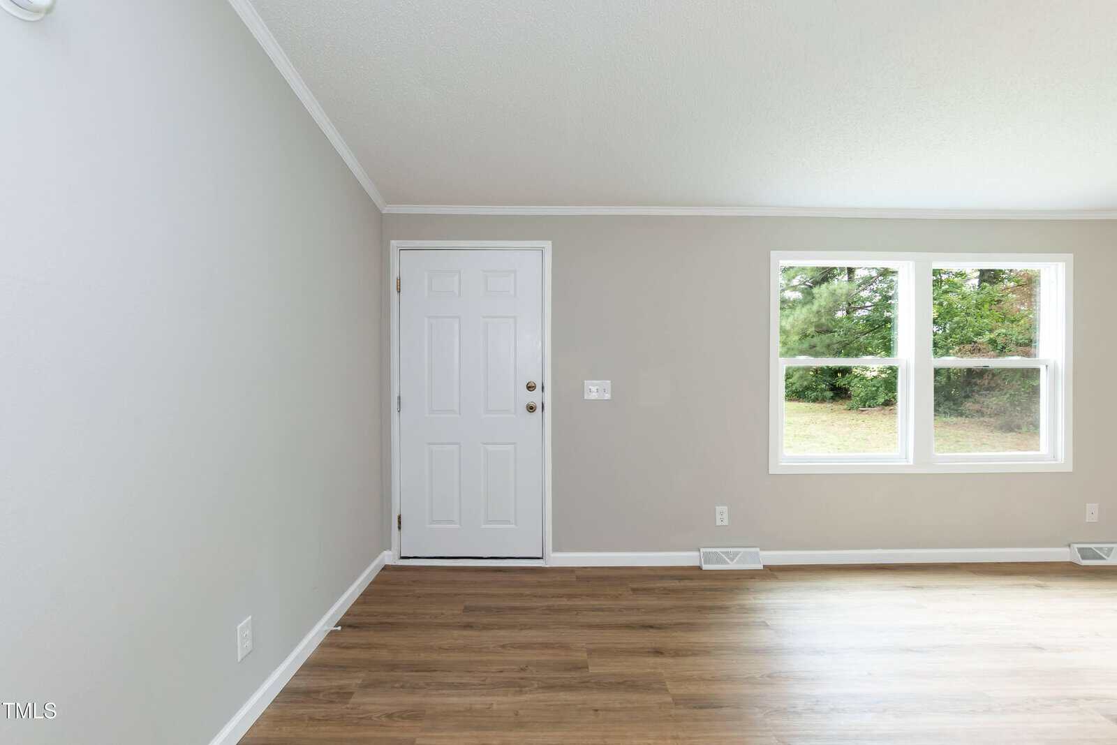 A room with light gray walls, white trim, wood flooring, a white door on the left wall, and two rectangular windows on the right wall showing greenery outside.
