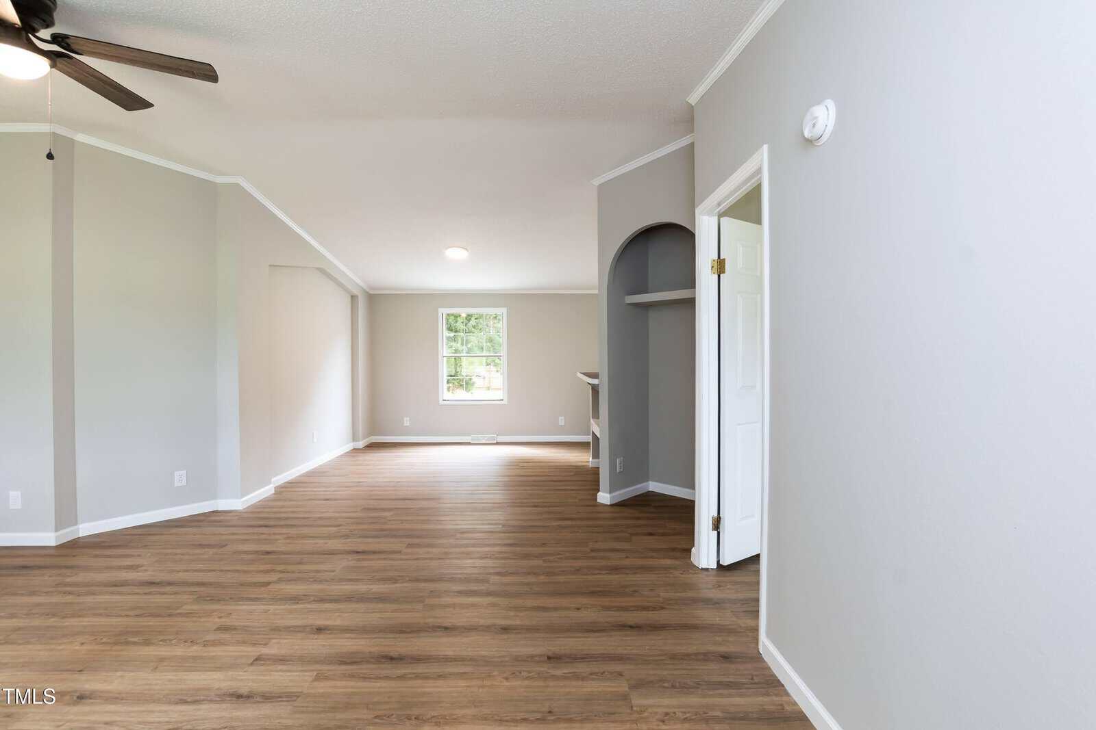 A spacious, empty room with light gray walls, wood-style flooring, white trim, and a ceiling fan. An arched doorway and an open white door are visible, along with a window letting in natural light.