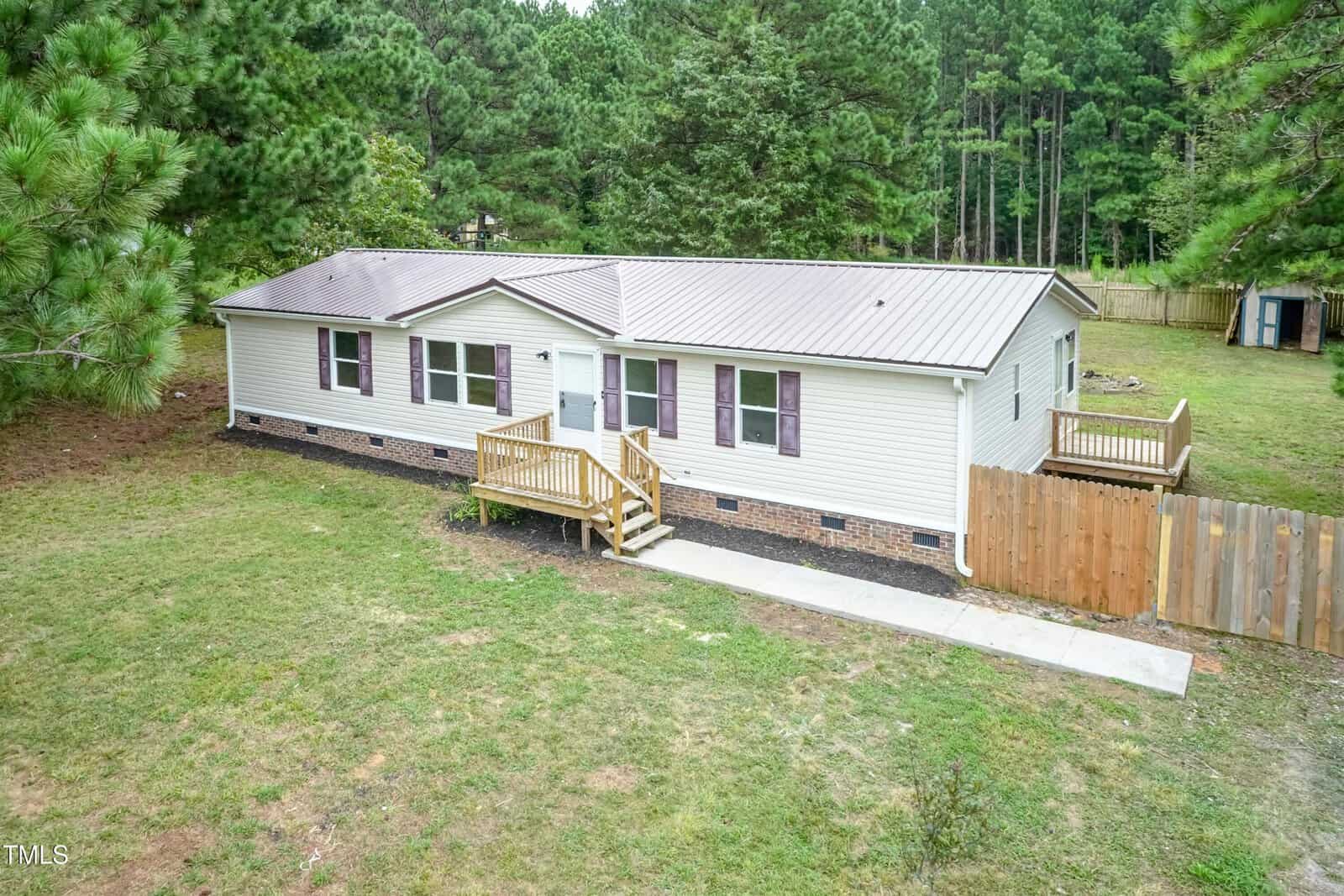 A single-story manufactured home with beige siding, a brown metal roof, a small wooden front porch, and a fenced backyard, surrounded by grass and trees.