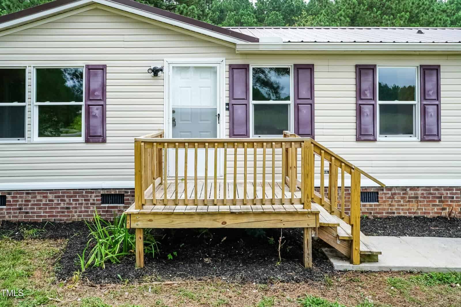 A beige house with purple shutters features a small wooden porch with steps and a railing leading to a white door. The porch is built over a mulched area with some plants. Brick foundation is visible below the siding.