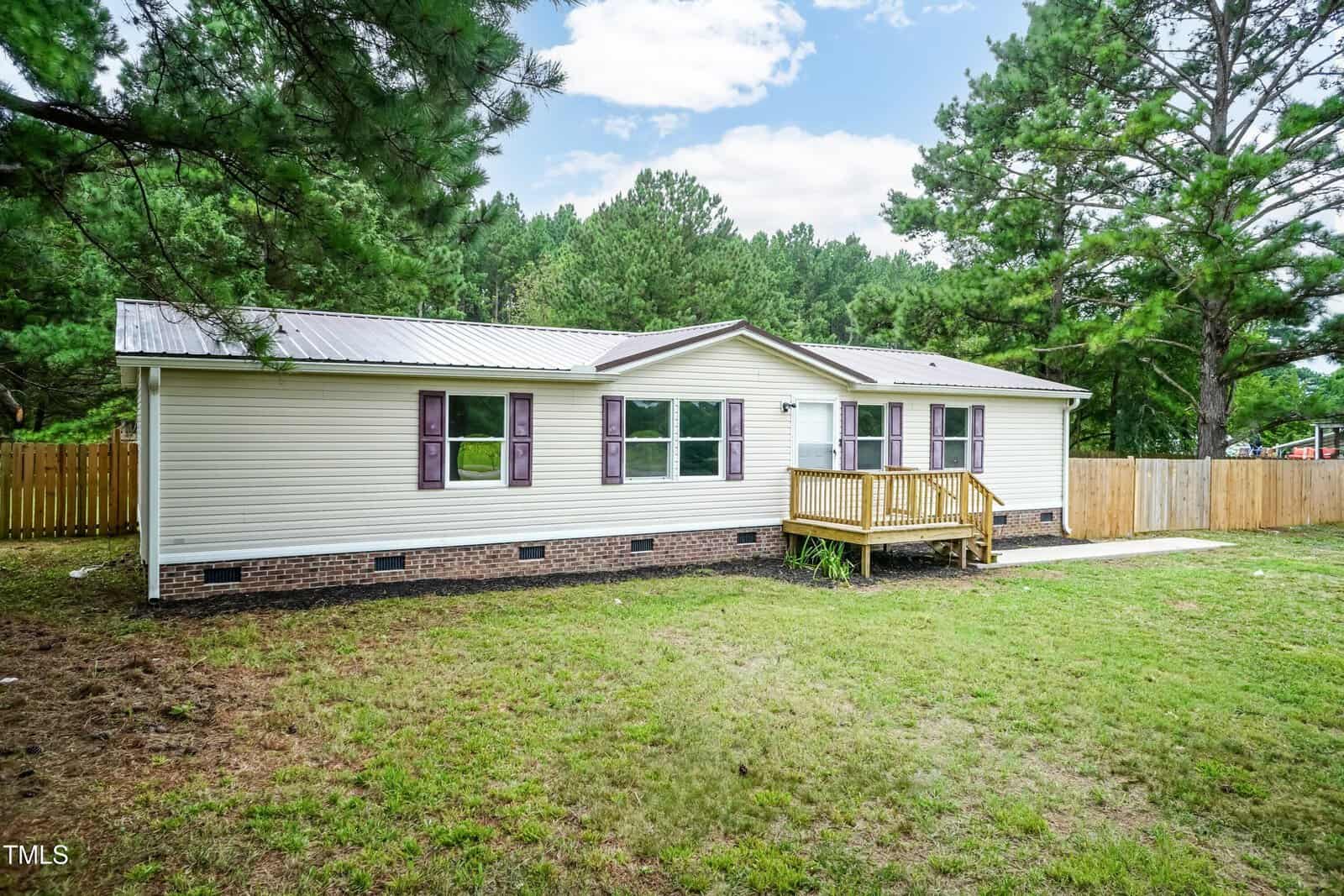 A single-story manufactured home with light-colored siding, a small wooden front porch, brick foundation, and purple shutters, set on a grassy yard with a wooden fence and surrounded by tall pine trees.