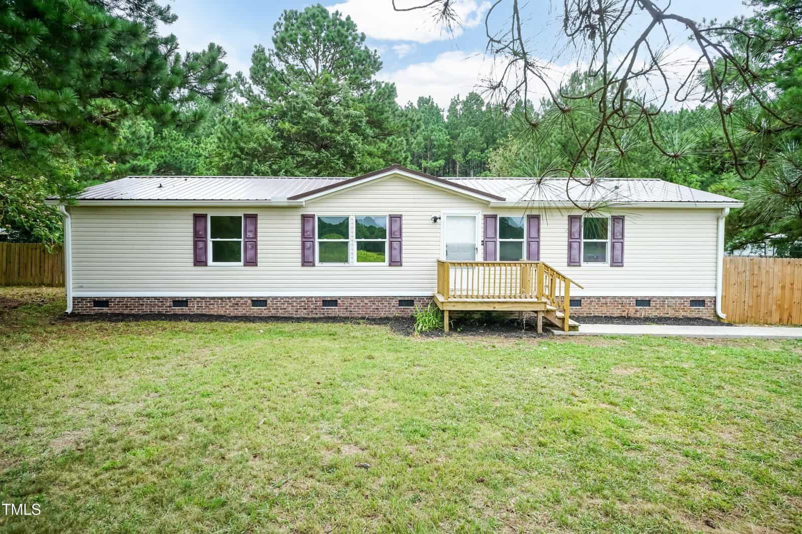 A single-story house with beige siding, a brick foundation, and a metal roof. The front features several windows with purple shutters, a small wooden porch with steps, and is surrounded by grass and trees.