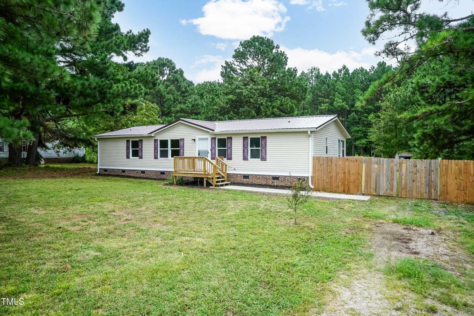 A single-story manufactured home with cream siding and purple shutters, surrounded by trees. The house has a small wooden porch, a fenced backyard, and sits on a grassy yard under a partly cloudy sky.