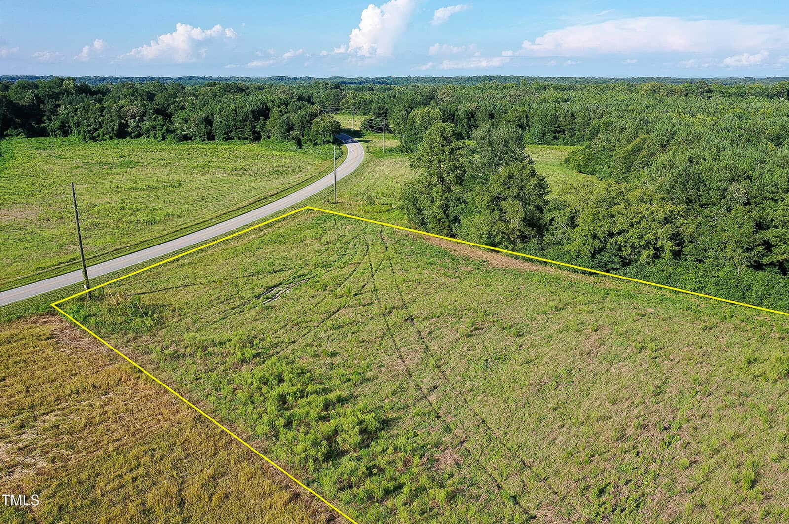 Aerial view of a grassy, vacant plot of land bordered by a yellow outline, next to a curving rural road and surrounded by green trees and fields under a partly cloudy sky.