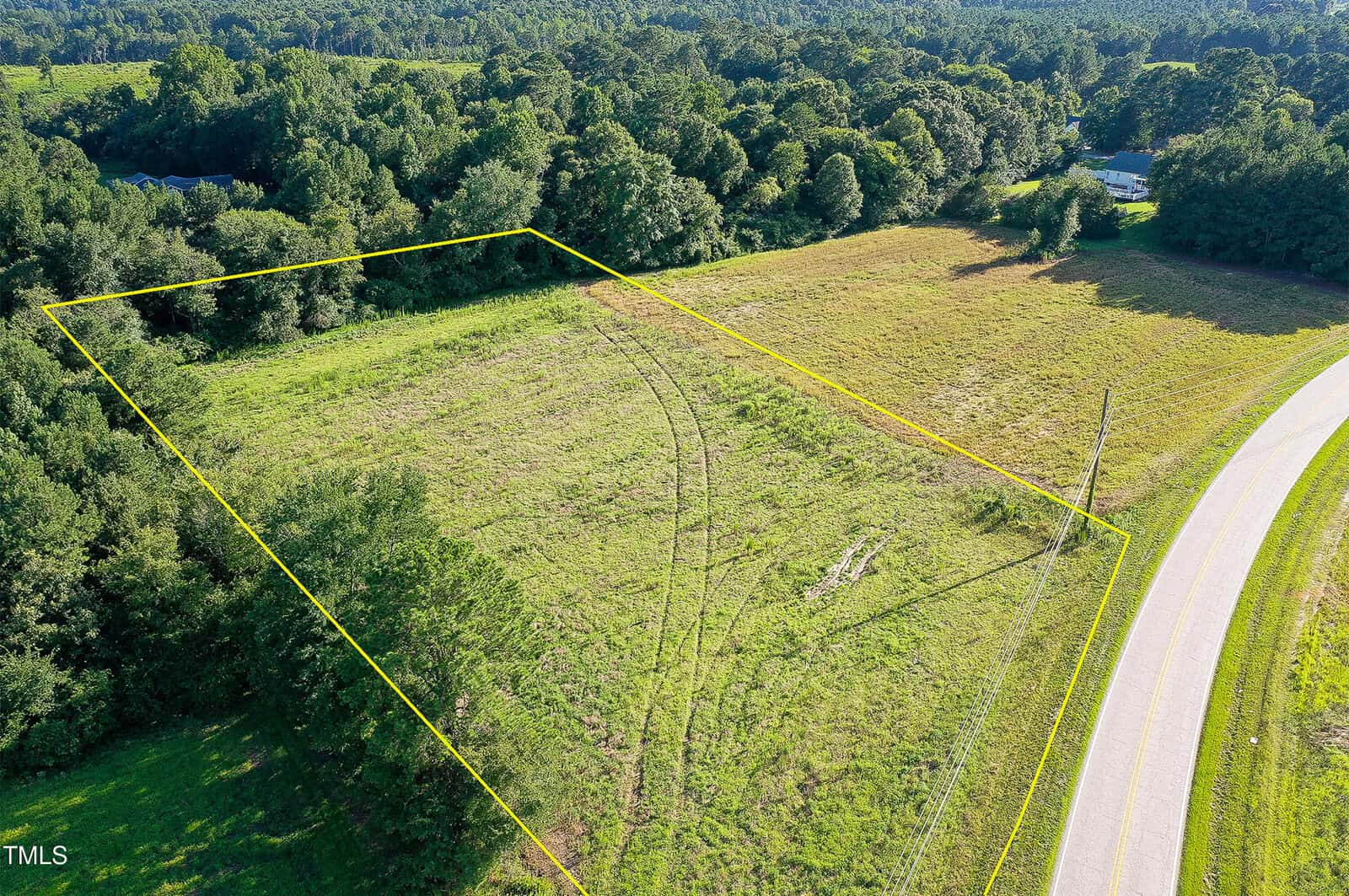 Aerial view of a grassy, partially wooded plot of land outlined in yellow near a curved road, with trees and forested areas surrounding the property.