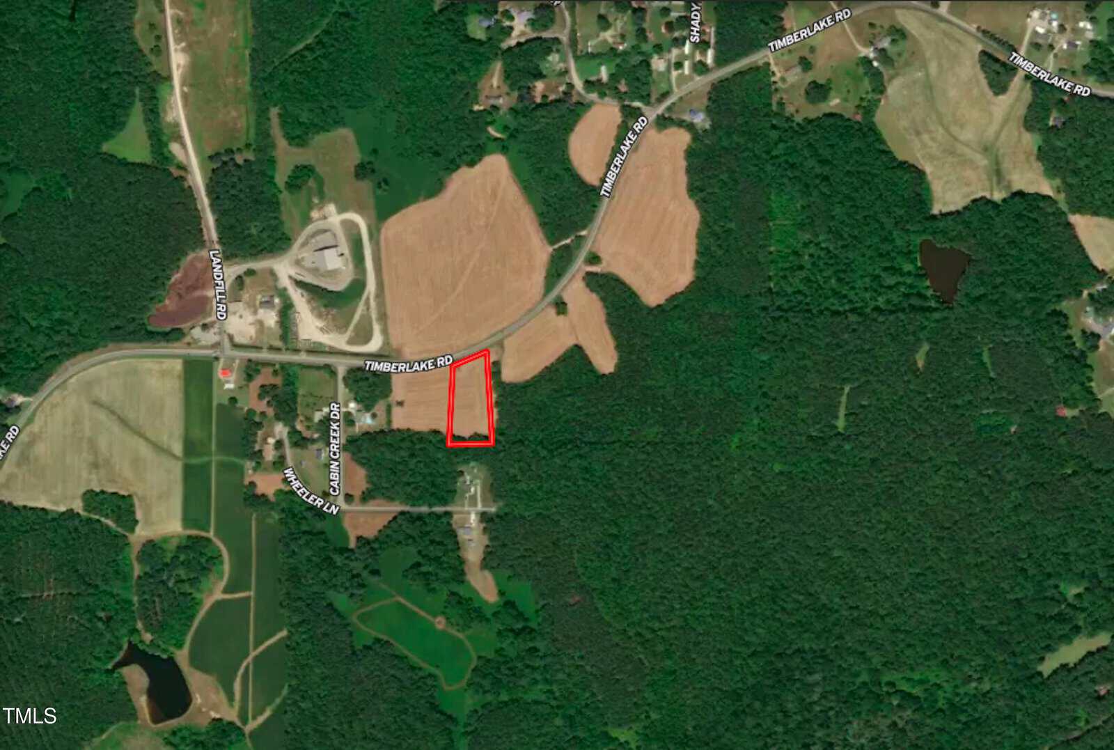 A satellite view of a rural area with forest, fields, and scattered houses. A rectangular plot of land is outlined in red near Timberlake Place road, surrounded by trees and farmland.
