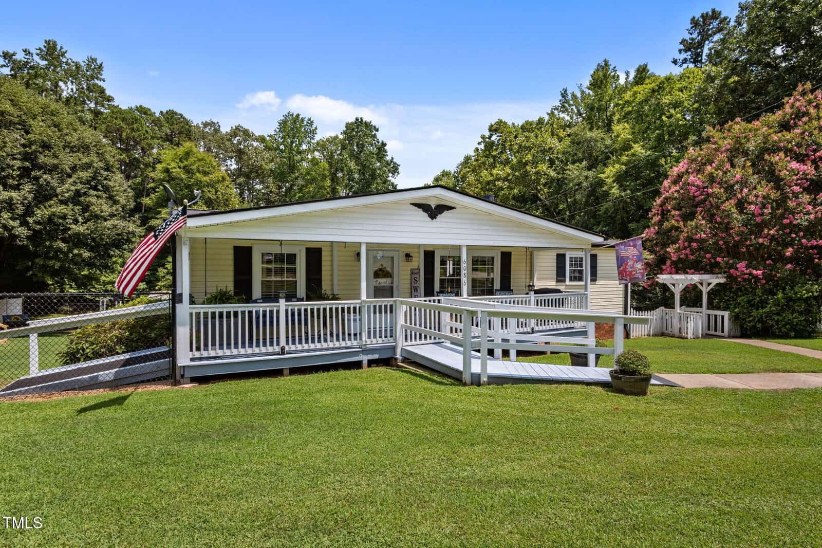 A single-story white house with a large front porch, wheelchair ramp, and American flag. Well-maintained lawn, trees, flowering shrubs, and a wooden arbor are visible under a clear blue sky.