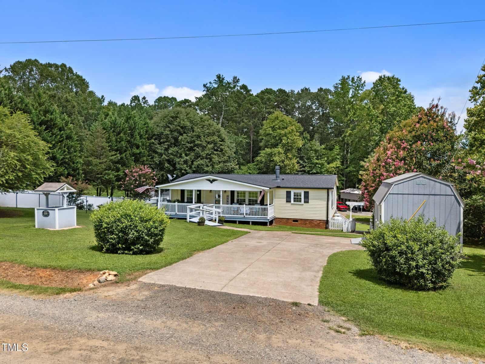 A single-story beige house with a covered front porch, a shed, and a small structure in the yard. The property features a concrete driveway and is surrounded by grass, trees, and shrubs under a clear blue sky.