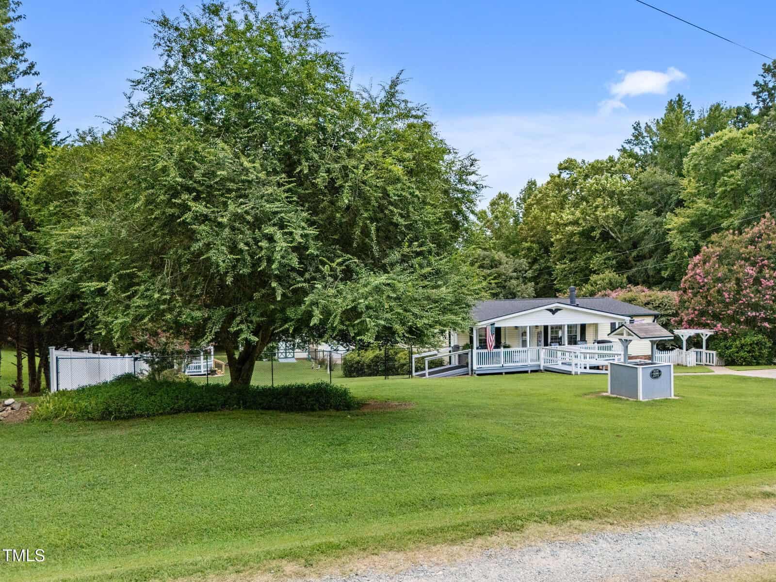 A small white house with a covered porch and ramp sits on a well-maintained grassy yard with large trees and shrubs. A white fence and a paved driveway are visible, with woods in the background.