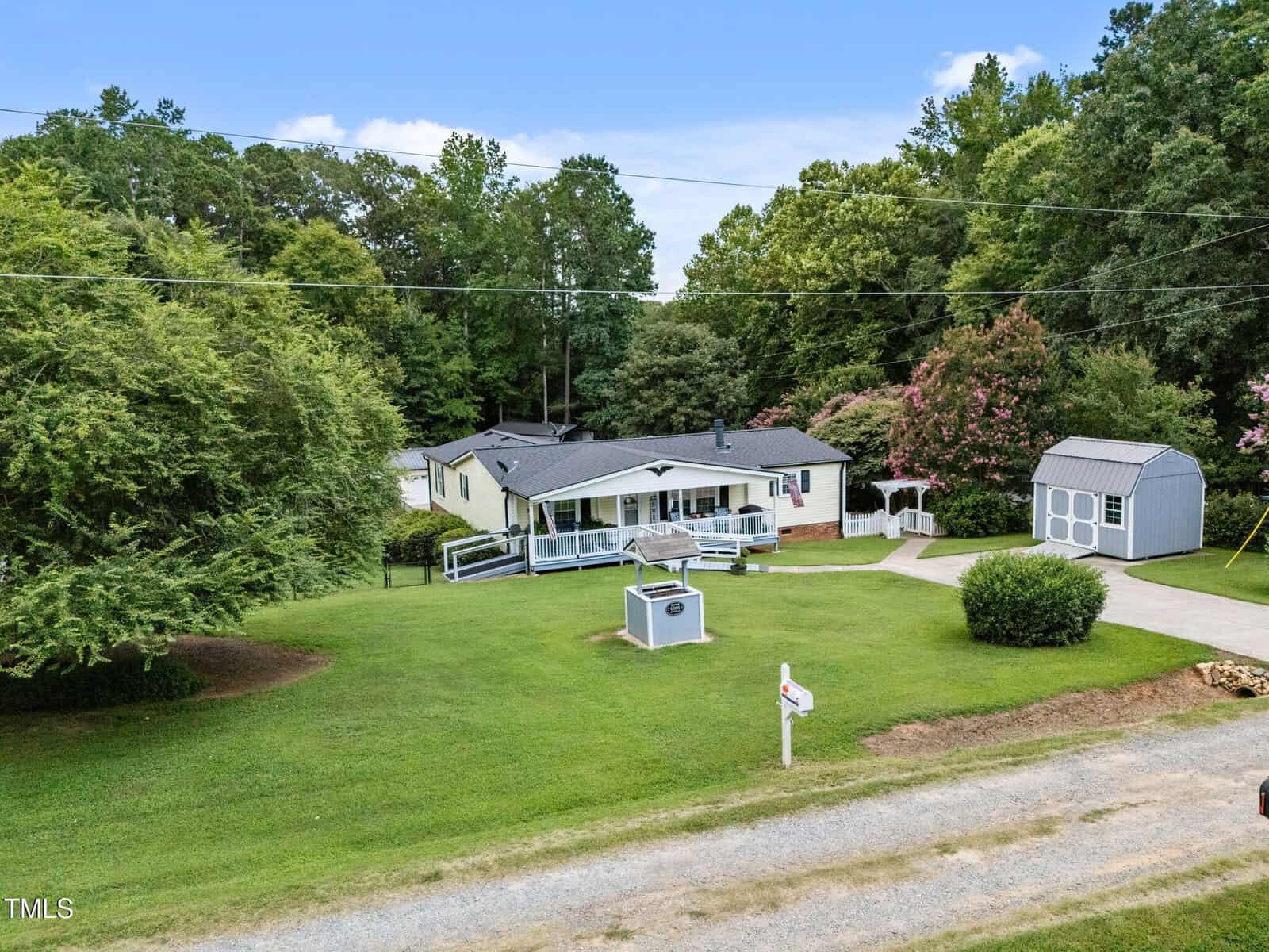 Single-story white house with a gray roof, wrap-around porch, and ramp. The home sits on a large lawn with a shed, trees, and a gravel driveway in a suburban setting. A mailbox and sign are near the road.