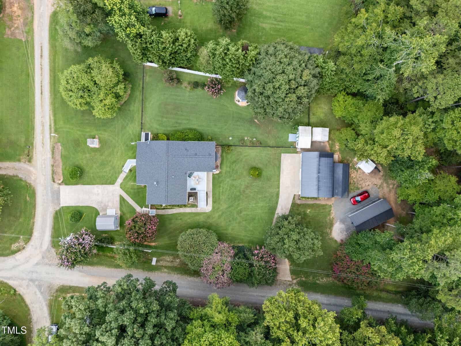 Aerial view of a property with a house, two detached garages, several trees, a large green lawn, and driveways connecting to a road. Multiple vehicles are parked on the property and in the driveway.