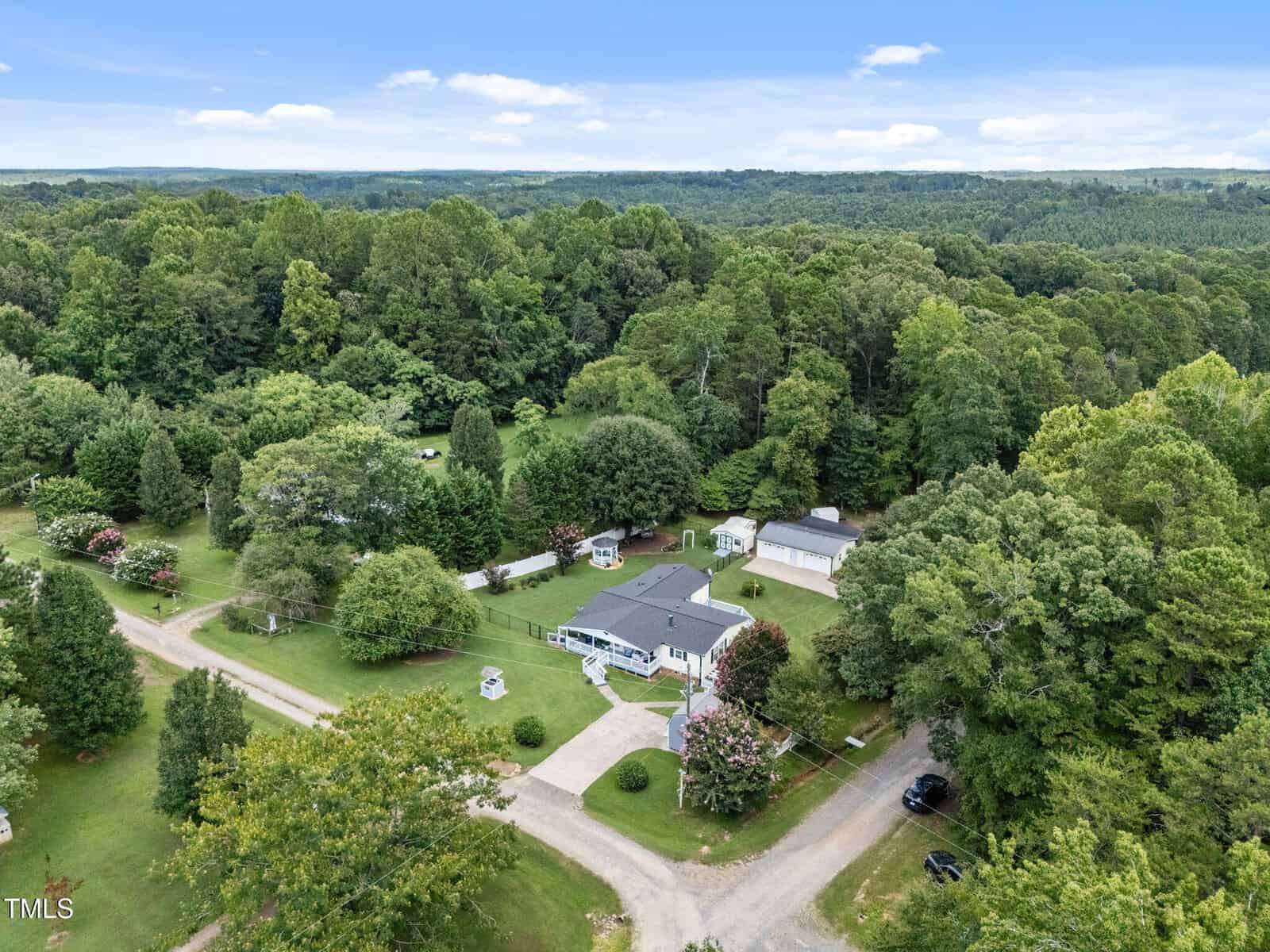 Aerial view of a house with a grey roof, large driveway, and surrounding lawn, set among dense trees and greenery, with multiple smaller buildings and a paved road leading to the property.