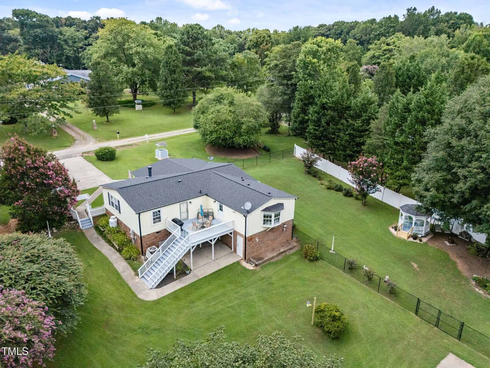 A light-colored, single-story house with a dark roof sits on a large, grassy lot with trees and gardens. A white staircase leads to an upper deck, and a gazebo is visible in the fenced backyard area.