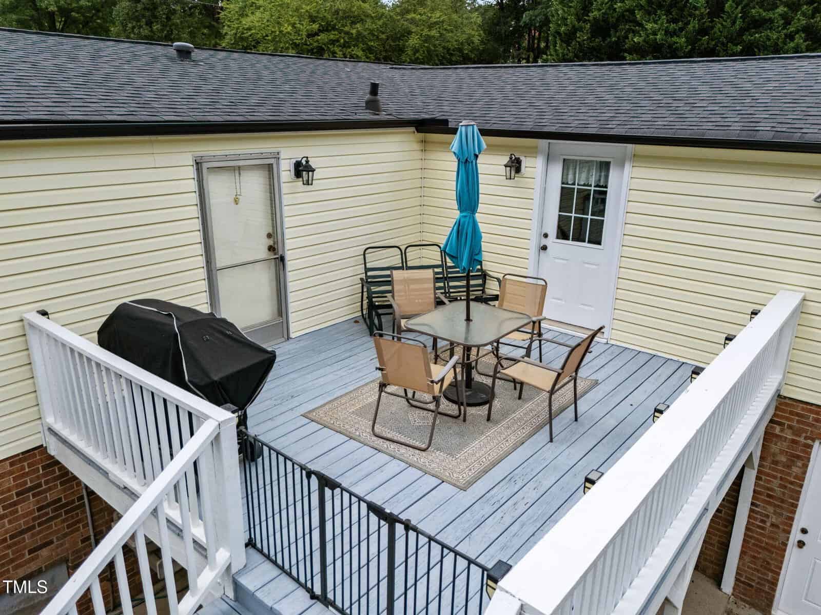 A raised outdoor deck with a light blue floor features a glass table, six chairs, a closed blue umbrella, a small rug, a grill, and two doors leading to the house. White railings enclose the space.