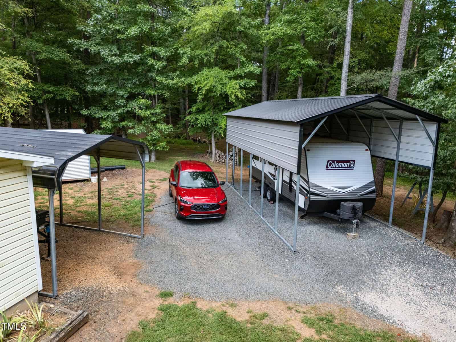 A red SUV is parked on gravel between a covered carport and a larger RV shelter with a Coleman travel trailer underneath. Trees and woods are in the background, and part of a light-colored building is visible on the left.