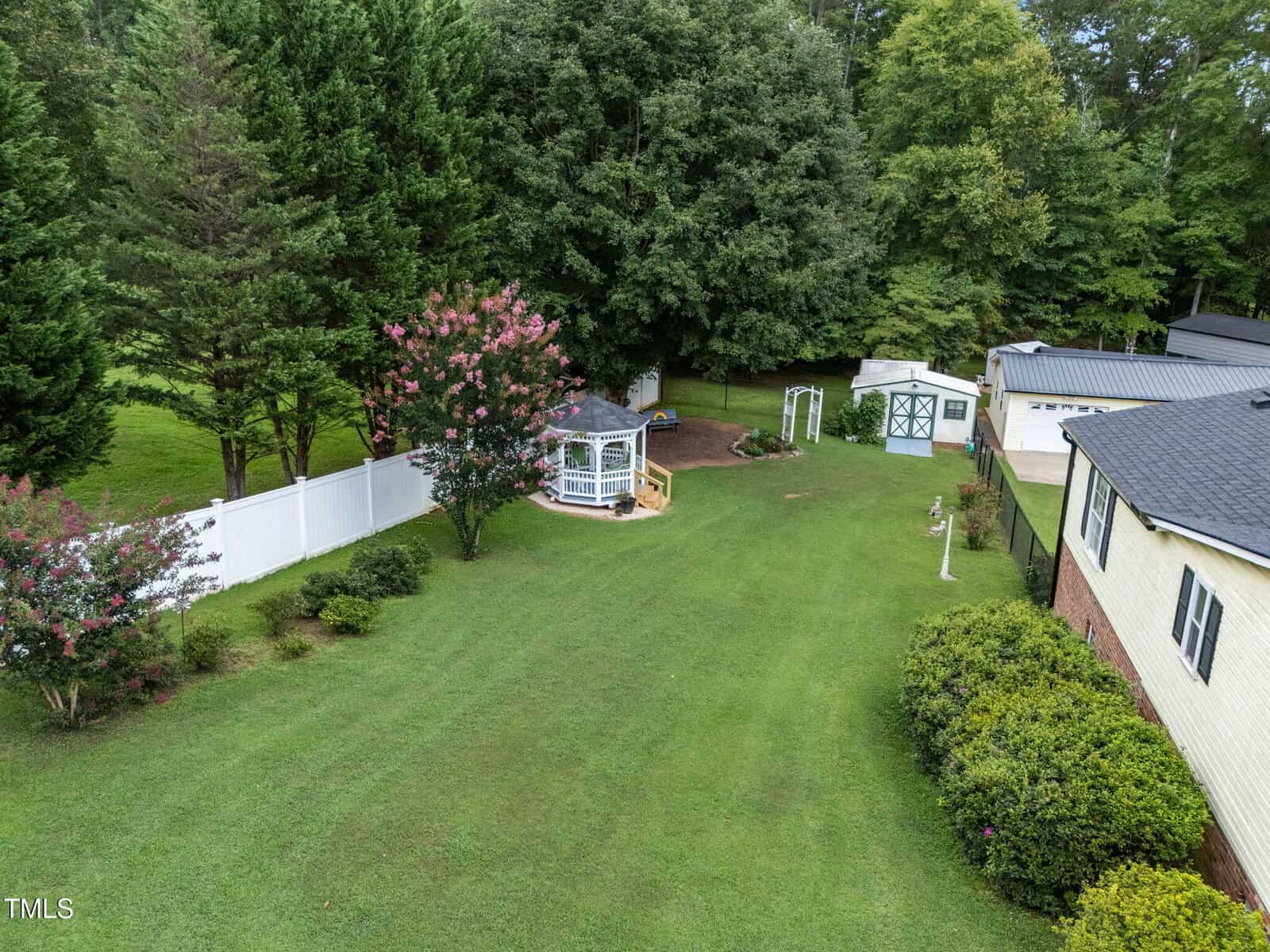 A spacious backyard with green grass, white fence, flowering trees, a white gazebo, shed, and part of a beige house with a dark roof, all bordered by tall trees in the background.